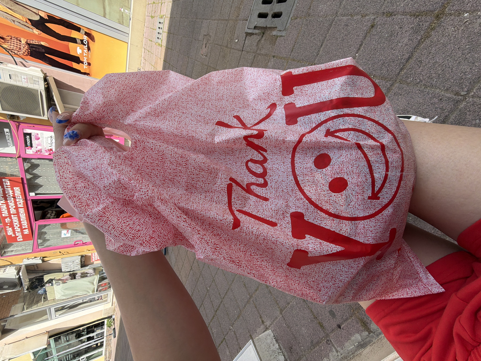 The image captures a first-person perspective of a person holding a red and white shopping bag on a sunny day in an urban setting. The prominent bag features the phrase "Thank YOU" in large red lettering, alongside a red smiley face logo that incorporates a recycling symbol. The person's arms are visible, holding the bag, and their legs, clad in red shorts, are also in the frame. Their fingernails are painted with a light base and a distinct blue design.
The background reveals a bustling street environment with grey block pavement and various commercial buildings. Bright sunlight casts clear shadows, indicating a sunny day. Storefronts are visible, displaying air conditioning units and advertising signs. Notable text on the signs includes "TOM TAILOR" on a poster, and several in Bulgarian: "БЪЛГАРСКИЯТ ПРОИЗВОДИТЕЛ НА ХАВЛИЕНИ ИЗДЕЛИЯ" (The Bulgarian manufacturer of towel products) and "„ЯНА" - гр. ПАНАГЮРИЩЕ" ("YANA" - Panagyurishte). Additional visible brand names are "SELECTIVE," "matrix," and "Nook." The scene depicts a casual moment of shopping or carrying purchases on a vibrant street in Pazardzhik, Bulgaria.