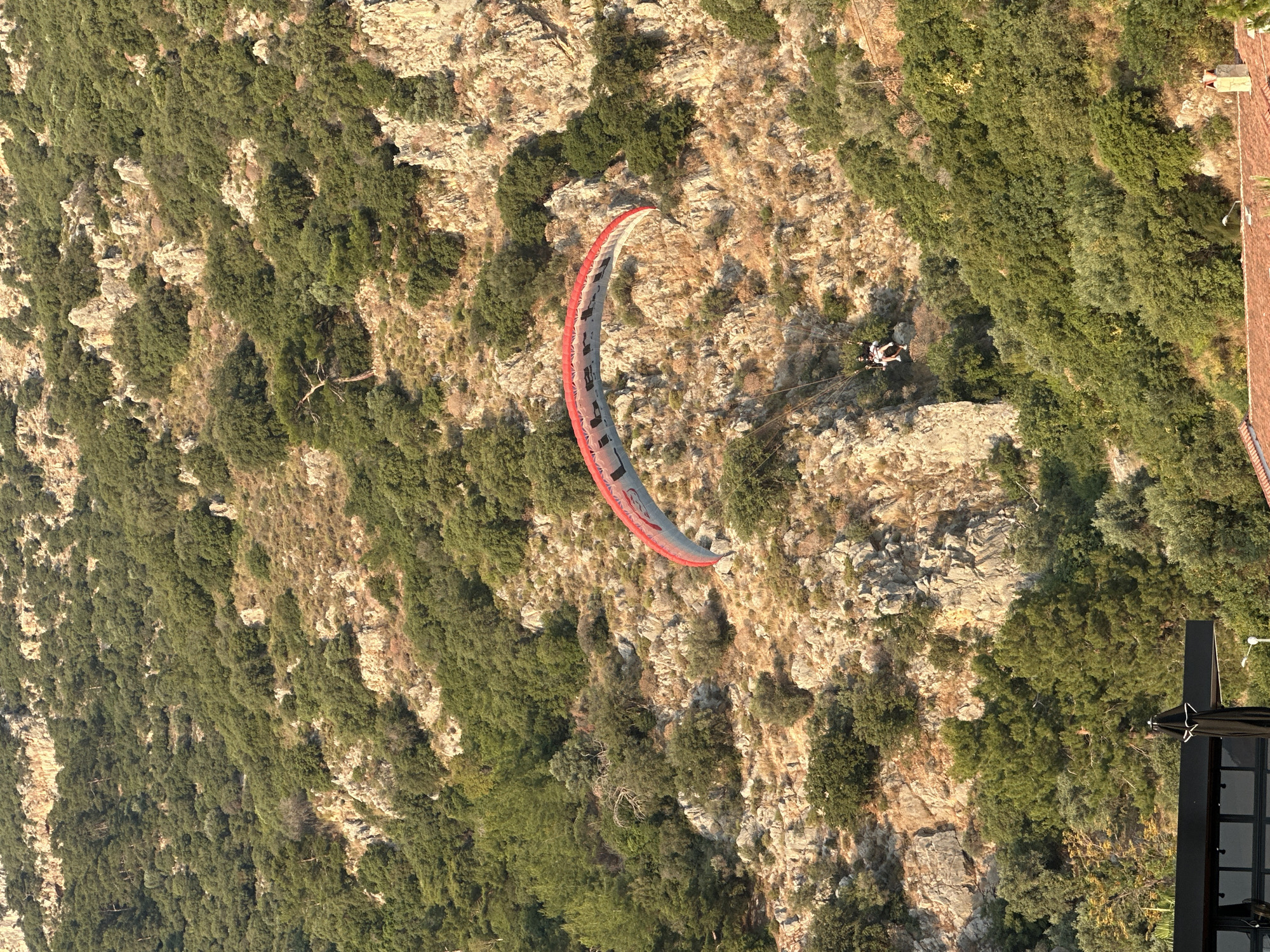 An image captures a paraglider descending along a steep, rugged mountainside in Fethiye, Turkey. The mountainside is densely covered with varying shades of green foliage, including numerous trees and shrubs, interspersed with prominent exposed rock formations that appear light brown or tan in the warm sunlight. Scattered patches of drier, lighter-colored vegetation are also visible on the slopes.
In the center of the frame, a paraglider canopy, predominantly red, white, and light blue/grey, is arced in flight. The word "Liberty" is discernable in white lettering on the light blue/grey section of the canopy. Suspended directly beneath the canopy are two individuals in a tandem harness, both appearing to wear dark clothing. They are positioned in a seated or reclining posture, indicating a controlled descent.
The upper portion of the image shows the peak of the mountain range, also covered in dense vegetation and rocks. In the lower foreground, parts of a structure are visible: a dark-colored, possibly metal, overhang with windows on the left, beneath which a closed black umbrella is positioned. To the right, a section of a tiled roof with red-brown tiles and a brick chimney is visible, indicating a building situated at the base or lower slope of the mountain. The lighting suggests either early morning or late afternoon, casting distinct shadows and highlighting the texture of the terrain.