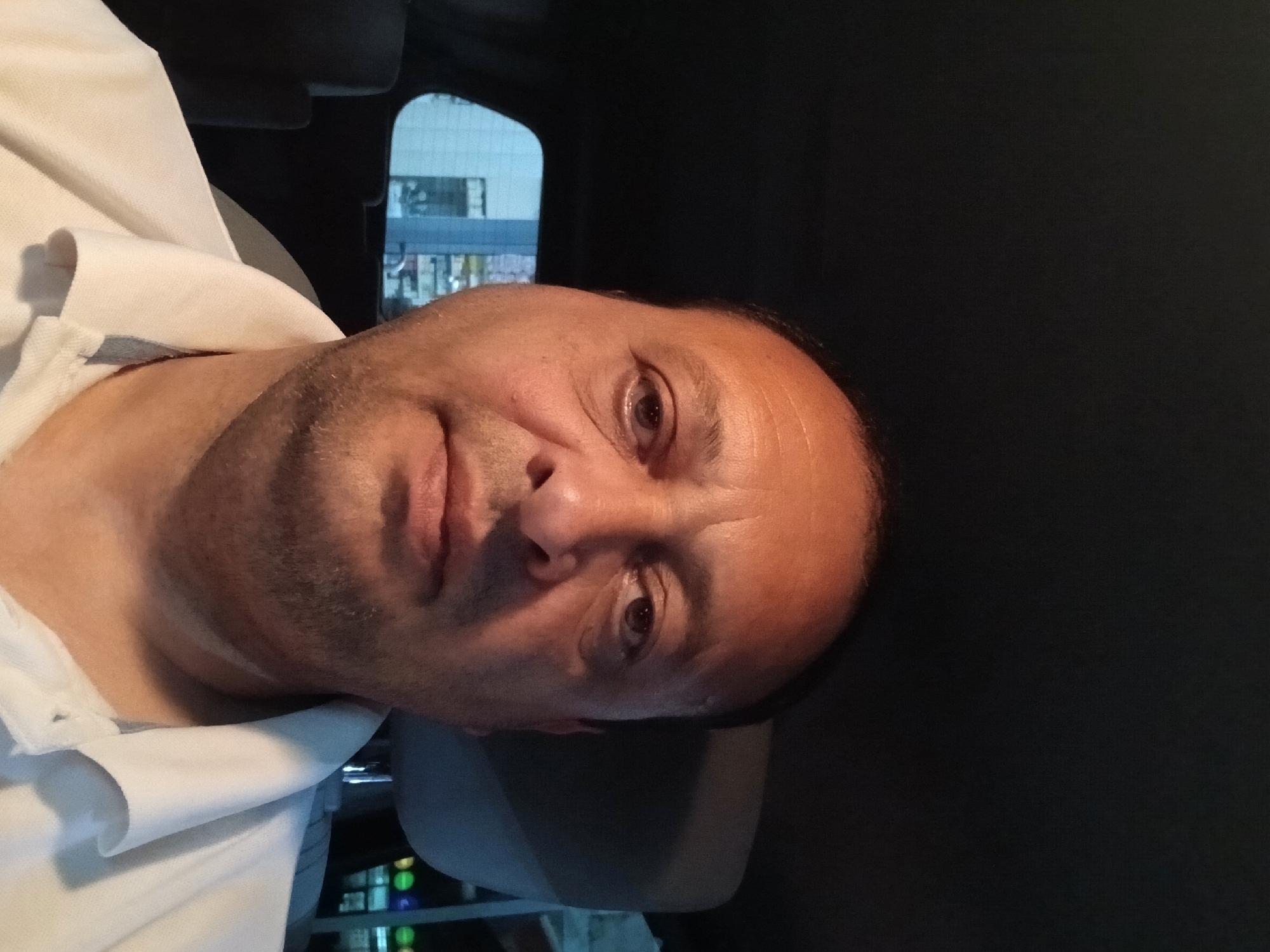 A man is pictured from the chest up, looking directly at the camera. He is wearing a white polo shirt with a grey collar. The background shows a car interior with a headrest and a window behind him. Through the window, buildings and streetlights are visible, suggesting an urban setting. The lighting suggests it is either dusk or nighttime. The location is identified as Telavi, Georgia.