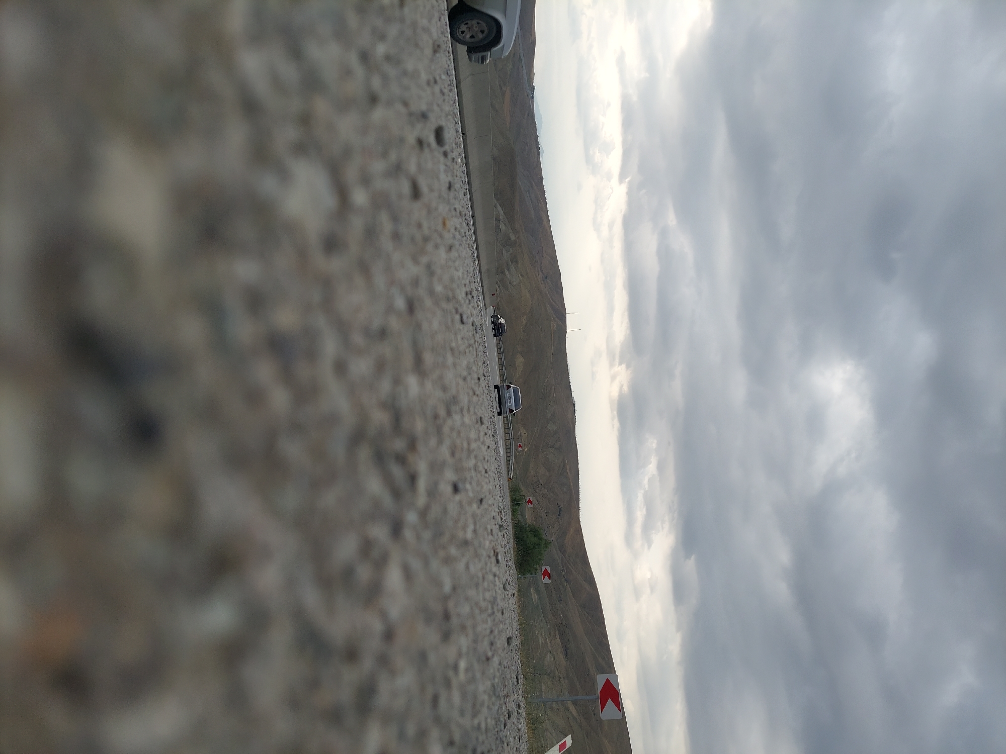 The image captures a low-angle view of a road with a gravelly surface in the foreground. In the middle ground, there is a paved road with several cars driving on it. The cars are mostly dark-colored, and their details are somewhat obscured by the distance and the perspective. To the left, a silver car is partially visible, also with its details softened by the angle and focus. Beyond the road, a concrete barrier runs parallel to the direction of travel. The background consists of rolling, arid hills under a cloudy, overcast sky. Several traffic signs with red and white chevrons indicating curves in the road are visible along the right side of the road. The overall impression is a highway scene in a relatively undeveloped, hilly landscape. The image appears to be taken in a rural or semi-rural area with a cloudy sky, suggesting potentially inclement weather or a general overcast day.