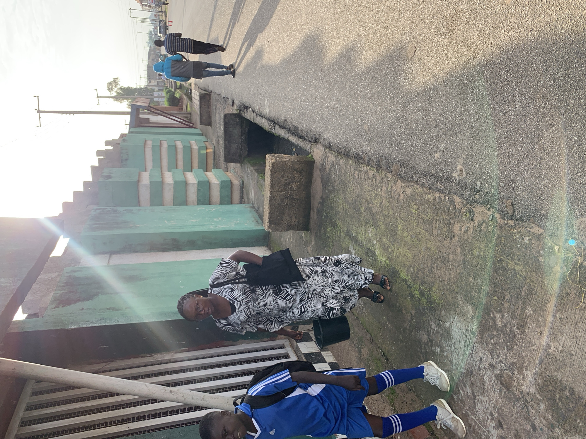 The image depicts a street scene in Jos, Nigeria, with a focus on a sidewalk alongside a road. In the foreground, a young boy in a blue soccer uniform with white striped socks and grey athletic shoes walks on the sidewalk, carrying a black backpack. Beside him, a woman in a black and white patterned dress and black sandals walks, holding a black bucket with her left hand and a black tote bag over her right shoulder. She is wearing glasses. Behind them, two young men are walking away from the camera on the road. One wears a blue hooded jacket and jeans, while the other wears a striped long-sleeved shirt and dark pants. The environment includes a green and white painted wall with a metal fence on the left, a concrete drain along the side of the road, and utility poles and trees in the background. The lighting suggests it is late afternoon, with the sun casting long shadows. The road surface appears to be asphalt.