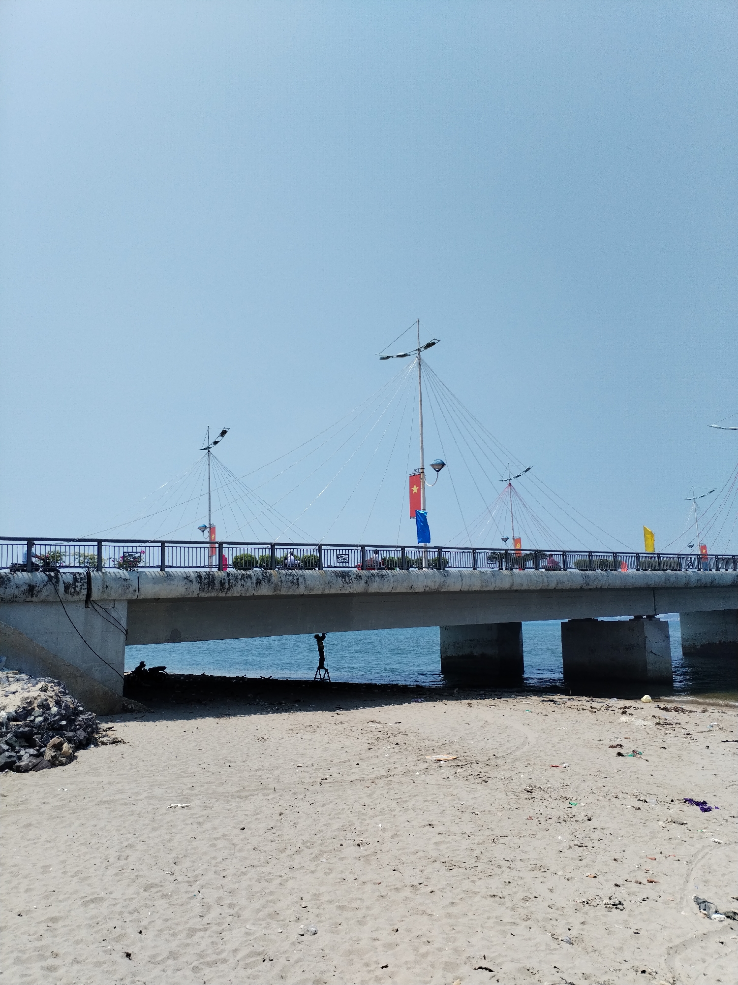 A bridge spans a sandy beach in Bac Nha Trang Ward, Vietnam. The bridge is supported by concrete pillars and adorned with flags and lights. A person stands on a stepladder beneath the bridge. Debris is scattered on the beach.