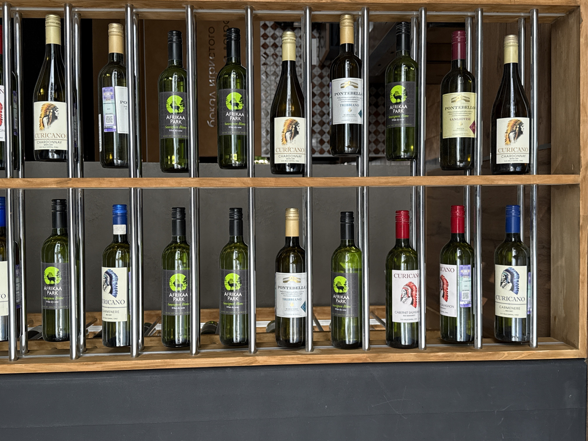 The image shows a wooden wine rack filled with bottles of wine. The rack is positioned against a dark grey surface. The wine bottles are arranged in vertical columns on multiple shelves. The bottles are of various types, including white and red wines, with labels indicating brands such as "Afrikaaz Park", "Curicano", and "Pontebello". The lighting suggests an indoor environment. The scene is static and appears to be a retail or storage setting. There are no people visible in the image. The specific location in Izhevsk, Russia, cannot be determined from the visual information provided.