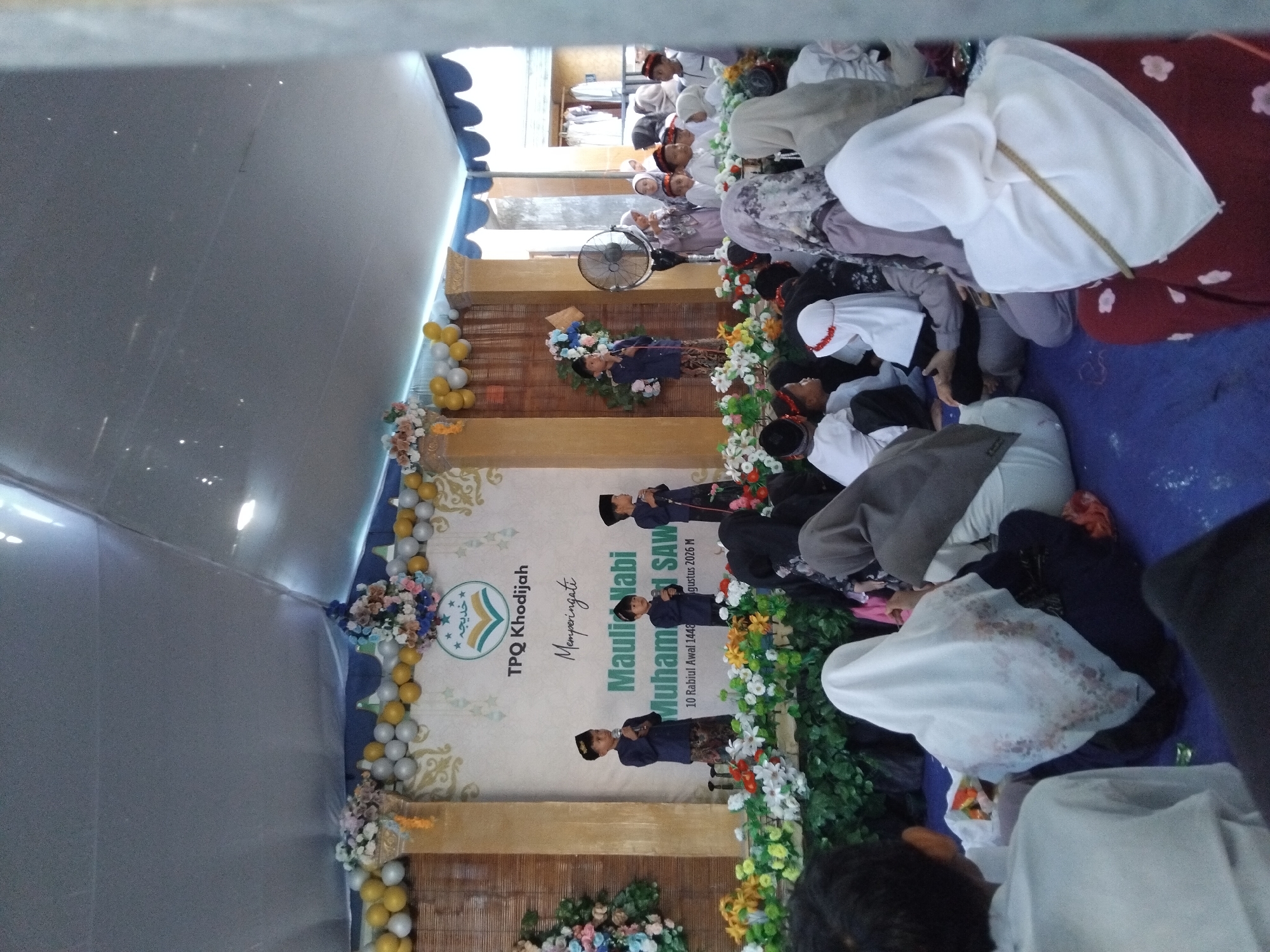 The image depicts an outdoor event, likely a religious celebration, taking place under a large tent in Kepuh Kiriman, Indonesia. The scene is filled with people, many of whom are children, dressed in Islamic attire. A group of young boys in blue shirts and patterned sarongs are standing on a stage, some holding microphones and appearing to perform. The stage is decorated with artificial flowers and balloons. A banner behind them reads "TPQ Khodijah Memperingati Maulid Nabi Muhammad SAW". In the foreground and extending to the sides are rows of attendees sitting on the ground, many of them women and children wearing hijabs. Some are looking towards the stage, while others appear to be engaged in their own conversations. The overall atmosphere seems to be one of community gathering and observance.