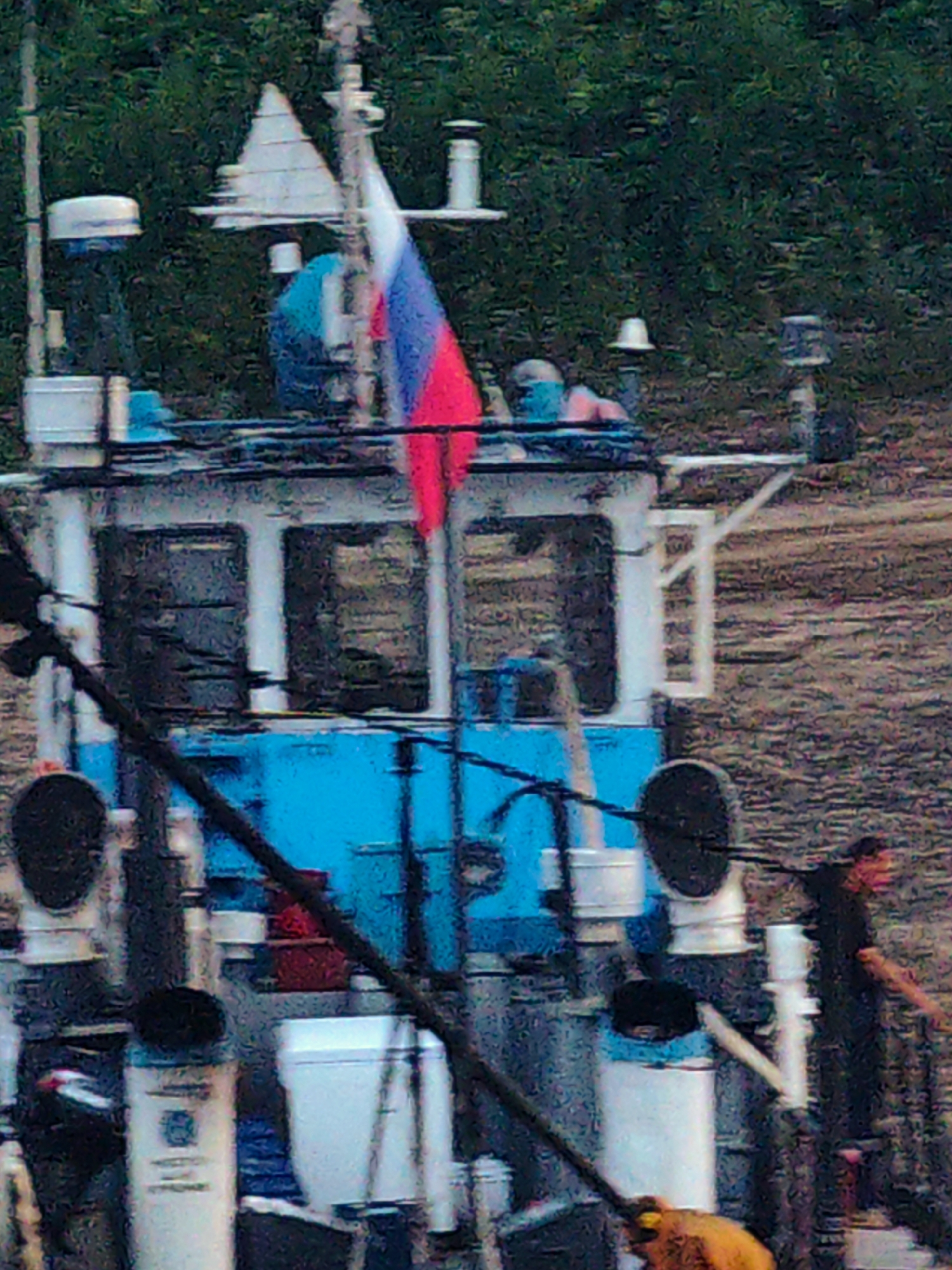The image shows a close-up view of a boat with a Russian flag flying. The boat appears to be docked or anchored, with trees and foliage visible in the background, suggesting a natural setting. The boat has a blue and white cabin with windows. Various pieces of equipment, possibly related to maritime operations, are visible on the deck, including pipes, cylindrical structures, and a black circular object.
A person in a dark shirt is standing on the right side of the boat, with their right arm extended, potentially interacting with something out of frame or signaling. Another person, partially visible at the bottom of the frame, is wearing an orange jacket. The scene appears to be during the day, with diffused lighting. The overall impression is one of a functional boat in a rural or natural environment. No text is visible in the image.