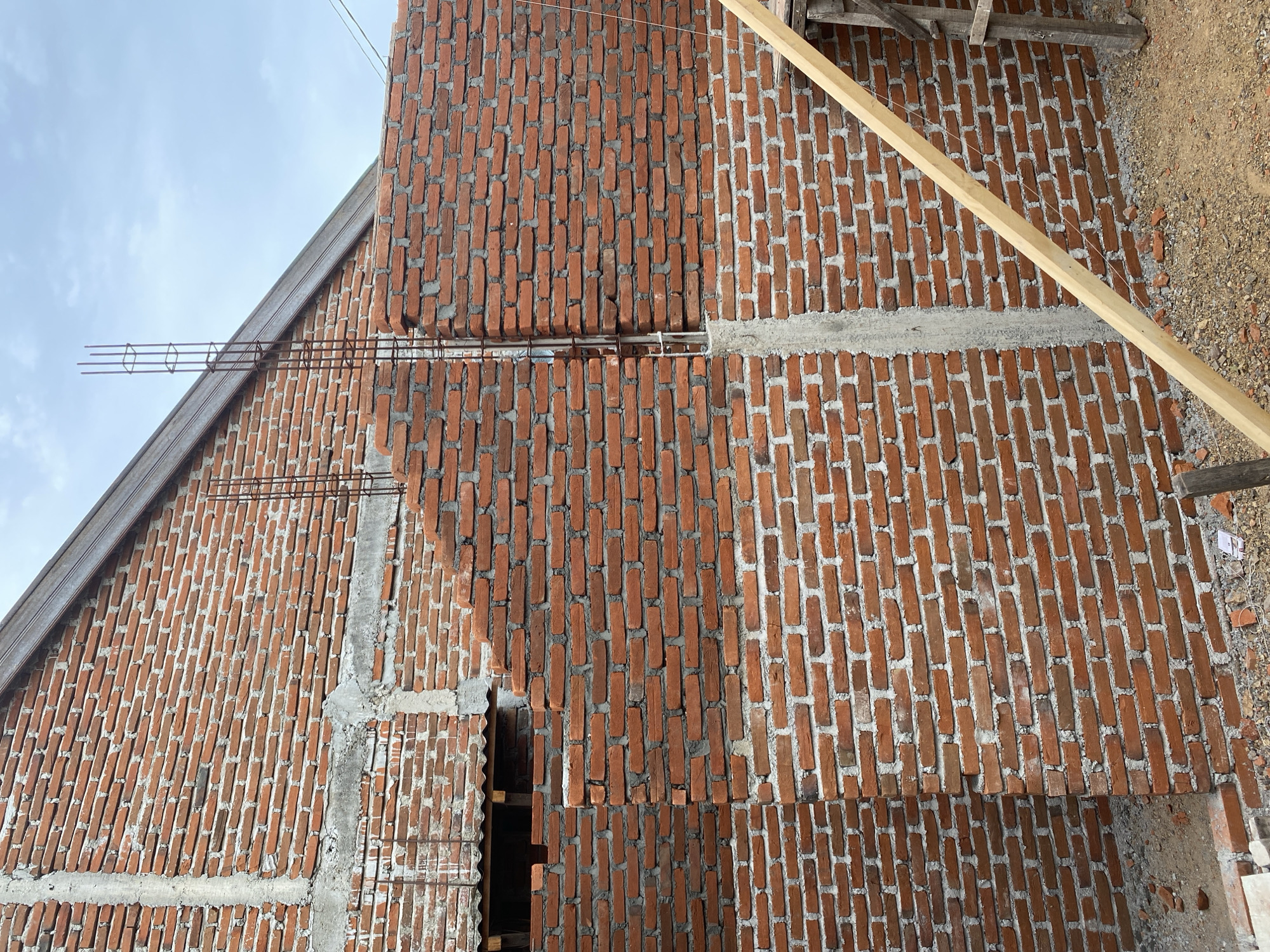 The image captures a construction site in Banda Aceh, Indonesia, focusing on an unfinished brick wall. The wall is constructed from red bricks, interspersed with concrete vertical supports and horizontal beams. Rebar cages are visible protruding from the top of some of these supports, indicating ongoing structural work. The foreground reveals a gravelly ground surface with loose bricks and construction debris. A wooden plank leans diagonally across the lower right portion of the frame, suggesting scaffolding or support structures. The sky above is partly cloudy. No people are present in the image. The overall scene depicts a stage of building construction in an urban or suburban environment.