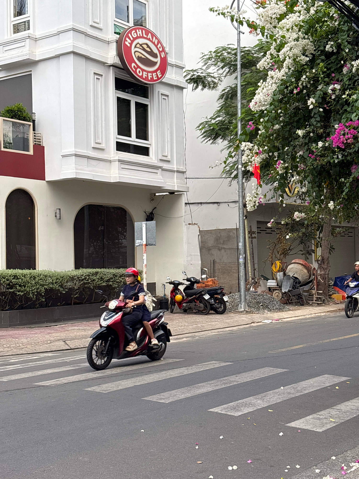 The image captures a street scene in Nha Trang, Vietnam, focusing on a pedestrian crossing and the surrounding urban environment. In the foreground, a woman wearing a blue dress and a red helmet is riding a dark red scooter across the white striped pedestrian crossing. She is looking forward, and a light-colored backpack is visible on her back. Behind her, another person on a white scooter is approaching the crossing from the right. The street is paved with asphalt, and scattered white and pink flower petals are visible on the road and the pedestrian crossing.
To the left of the street, a white building with decorative architectural elements stands prominently. A circular red sign with white lettering that reads "HIGHLANDS COFFEE" is mounted on the building's facade. Below the sign, a rectangular window is visible. A hedge runs along the base of the building.
To the right of the street, a sidewalk is visible, with several motorcycles parked. Further back, there is a construction area with a cement mixer, piles of gravel, bricks, and construction materials. Trees with white and pink flowers are growing alongside the sidewalk, with some branches extending over the street. The sky appears overcast. The overall scene depicts a typical streetscape in a Vietnamese city, with a blend of commercial establishments, residential buildings, traffic, and ongoing construction.