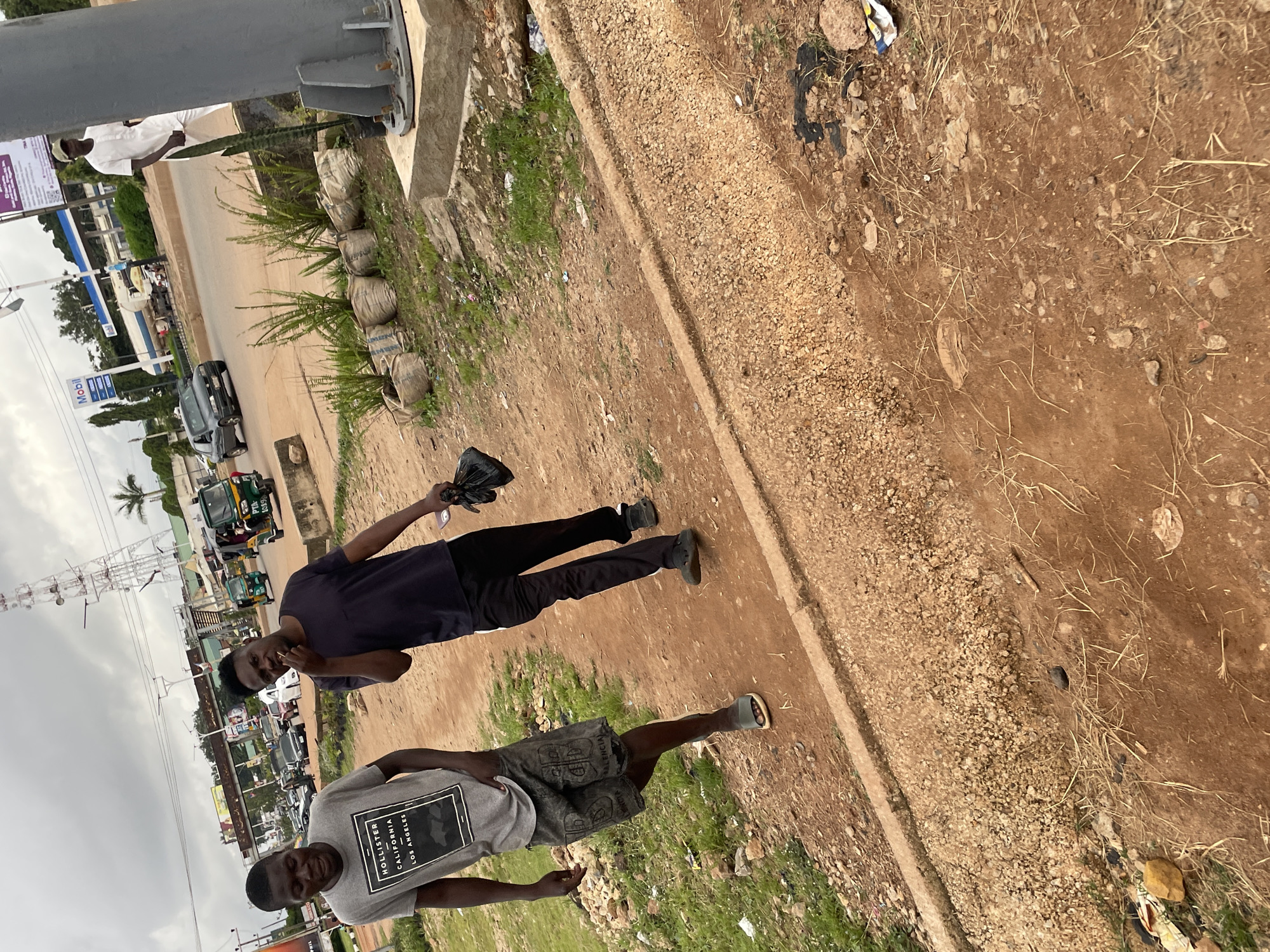 In Jos, Nigeria, two young men walk side-by-side on a dirt path adjacent to a busy road. The man on the left wears a grey t-shirt with the text "HOLLISTER CALIFORNIA LOS ANGELES" and patterned denim shorts. The man on the right wears a dark t-shirt, black pants with a white stripe, and holds a black plastic bag in his right hand while gesturing with his left. Behind them, a street is filled with traffic including cars, motorcycles, and a tricycle-like vehicle. The environment is characterized by a cloudy sky, sparse green vegetation, and the presence of various objects like plastic bags, debris, and potted plants along the roadside. A tall communication tower is visible in the distance.