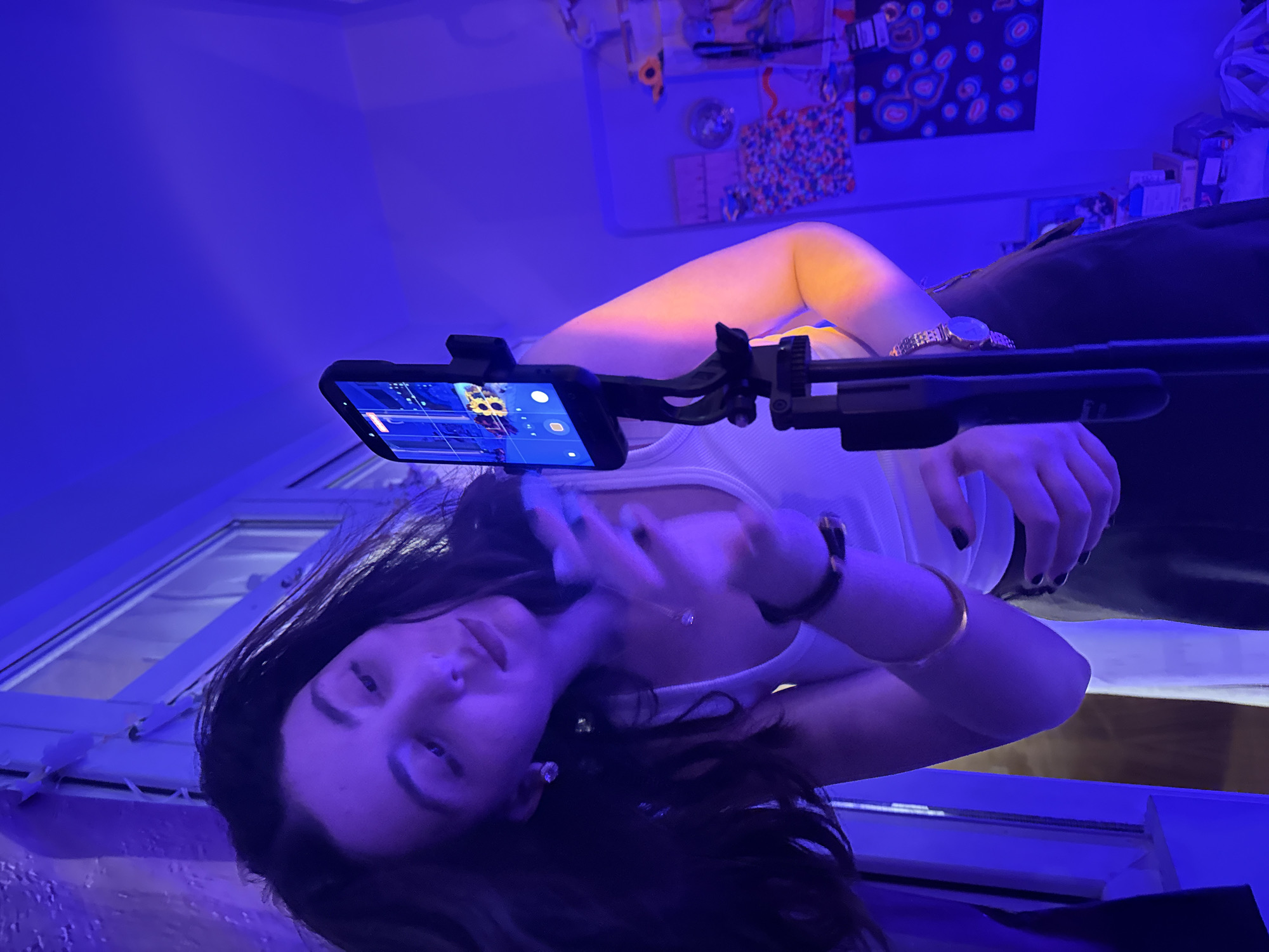 A woman is positioned in front of a mobile phone mounted on a tripod. She is wearing a white top and has dark hair. She is facing the camera with her right hand raised toward her face, possibly holding a smartphone. The environment is a room illuminated with blue light, with a window visible in the background. The location appears to be indoors. The room contains various objects, including artwork on the wall, and general clutter that indicates a lived-in space. There is no clear indication this photo was taken in Yerevan, Armenia.