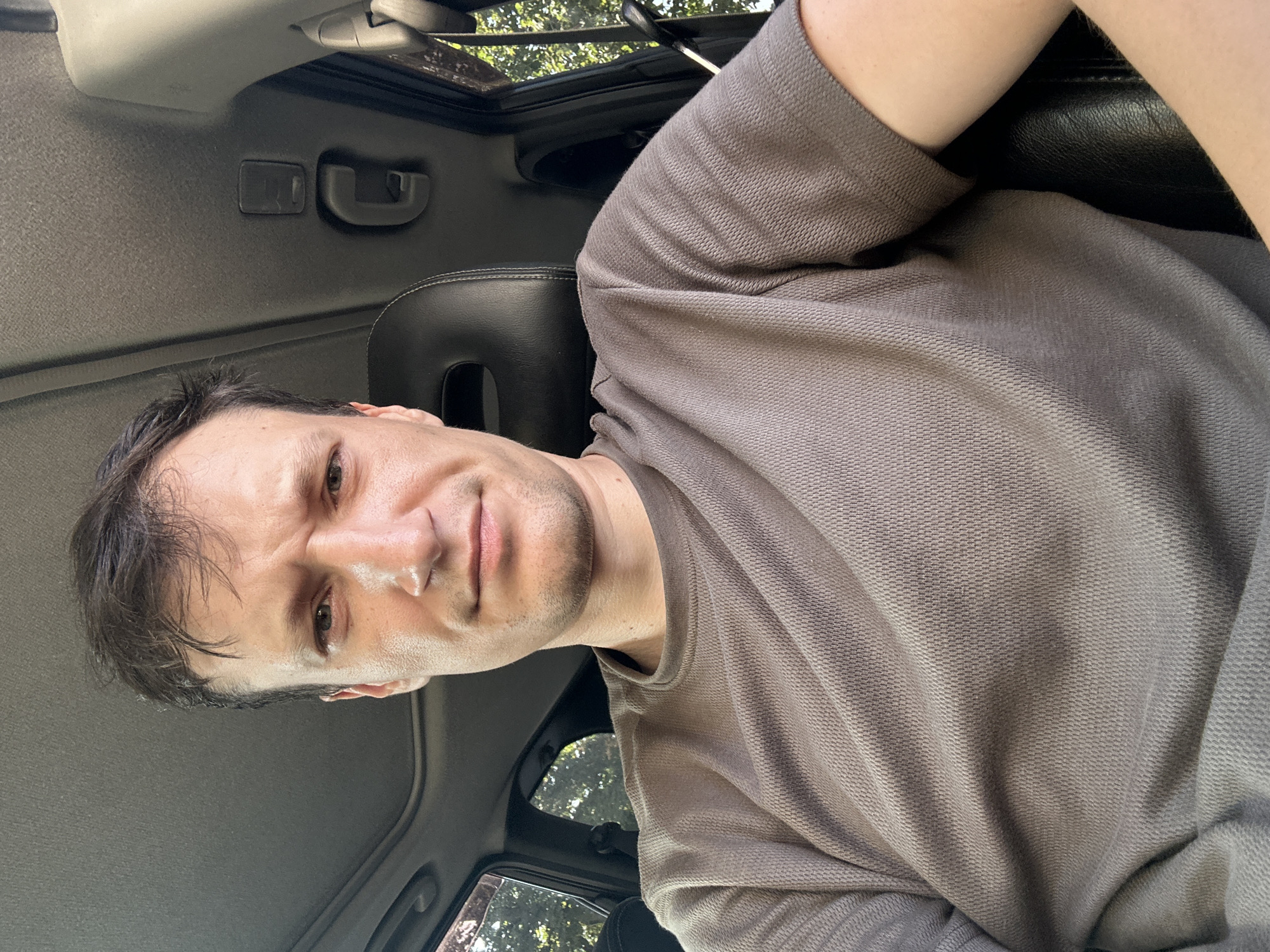 A man is sitting in the driver's seat of a car. He is wearing a brown t-shirt and has dark hair. The man is looking at the camera with a neutral expression. The interior of the car is visible, including the dashboard, steering wheel, and seats. The exterior of the car is partially visible, showing trees and foliage. The image appears to have been taken during the daytime. The location is not specified in the image, but it is mentioned that the location is in the city of Kamianets-Podilskyi, Ukraine.