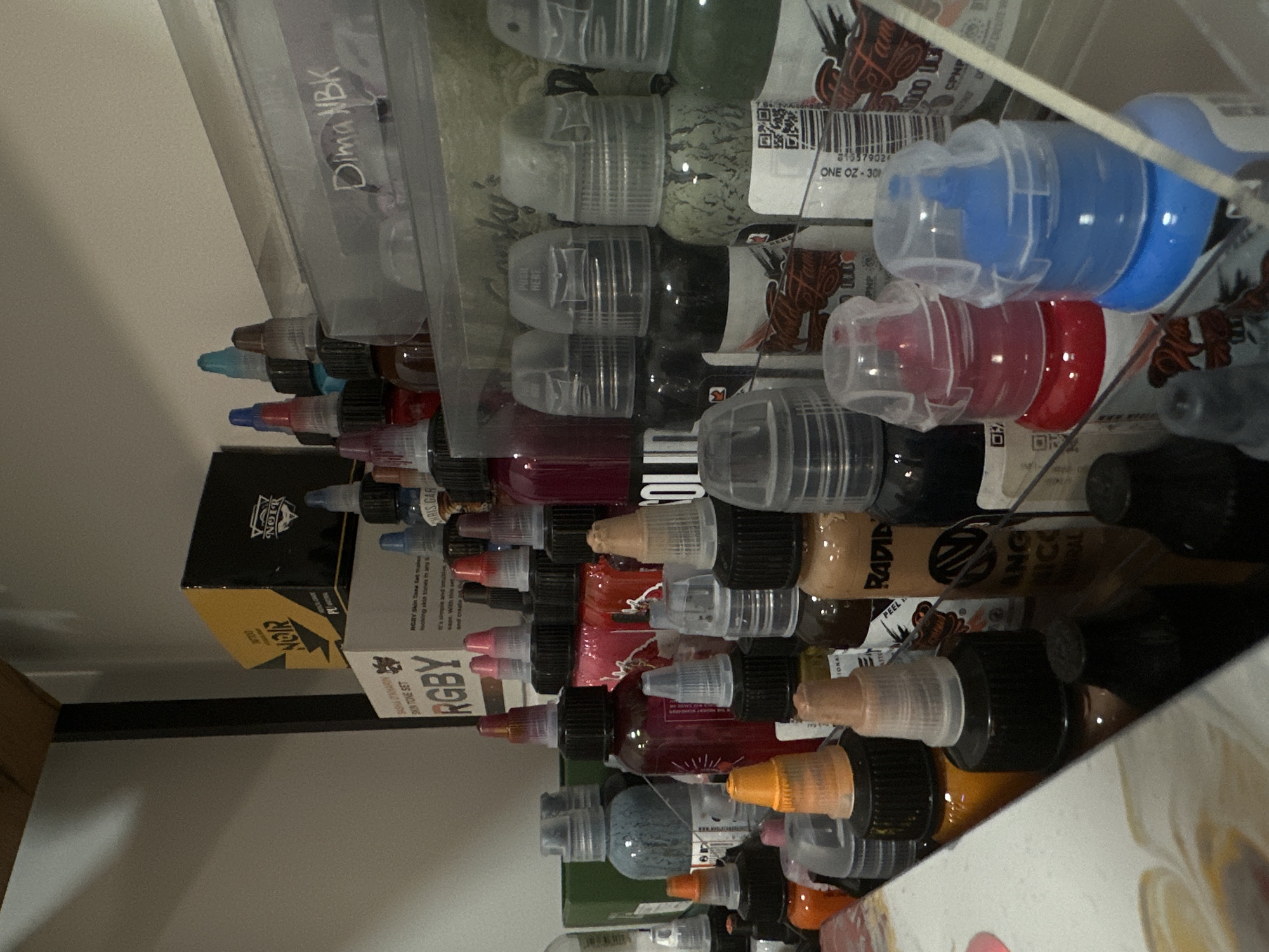 The image shows a shelf filled with bottles of tattoo ink, along with some product boxes. The brands visible include RGBY skin tone set, NOIR, Rapid, and other assorted brands. The bottles appear to be packed tightly together. A clear plastic container labeled "Dima NBK" sits above the ink bottles. The location in Rostov-on-Don, Russia, cannot be determined from the image.