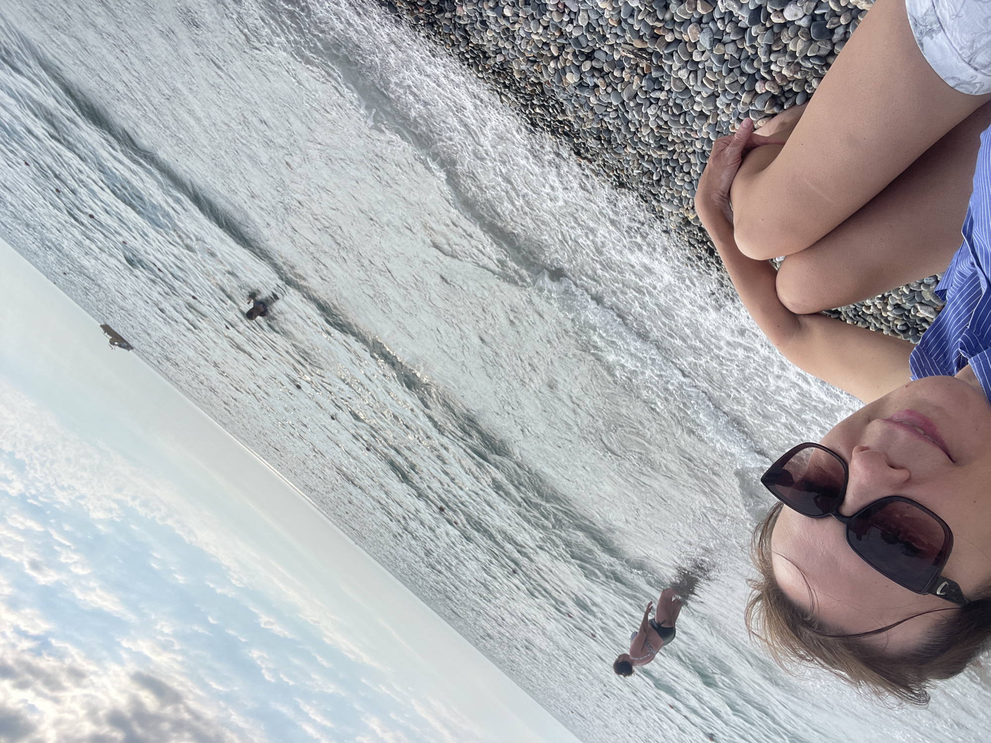 The image is a selfie taken at a beach in Sochi, Russia, depicting a woman sitting on a pebble beach with the sea in front of her. The woman is wearing sunglasses and a blue and white striped top, and she is looking towards the camera with a slight smile. Her legs are crossed, and she is resting her arms on her knees.
The sea is calm with gentle waves rolling onto the shore. In the distance, two people are visible in the water, one appearing to be swimming or wading, and another further out. The sky is partially cloudy, suggesting a pleasant day. The beach is covered in smooth, grey pebbles of various sizes. The overall mood is relaxed and summery. The time of day appears to be late afternoon or early evening, given the soft light. There is no visible text in the image.