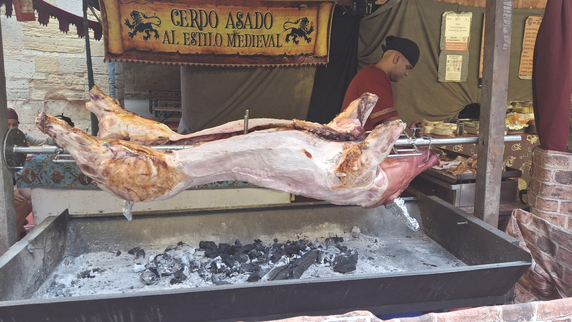 A whole pig is roasting on a spit over a charcoal grill. The pig is positioned horizontally, with its head facing left and its body extending to the right. The skin of the pig is partially browned, indicating it has been cooking for some time. Below the pig, a metal tray contains glowing charcoal embers and ash. A person wearing a red tunic and black headwear is visible on the right side of the image, tending to the spit. In the background, a banner with the text "CERDO ASADO AL ESTILO MEDIEVAL" (Roasted Pig in Medieval Style) is displayed. The setting appears to be an outdoor market or festival, suggested by the rustic wooden structures and brickwork visible. This scene is located in La Fresneda, Spain, a town known for its medieval festivals where such traditional cooking methods are often showcased.