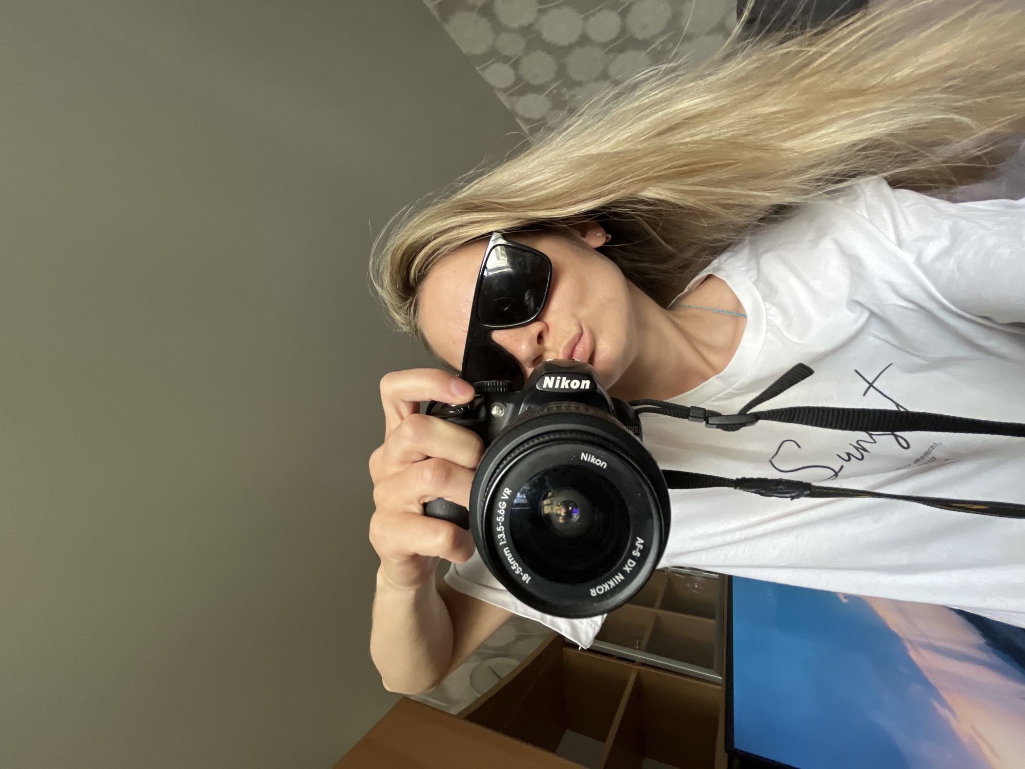 Here is a detailed description of the image:
A low-angle selfie of a blonde woman holding a Nikon camera. She is wearing sunglasses and a white t-shirt with the word "Sun" written on it. The camera has an 18-55mm lens attached. The background appears to be an indoor setting, with a grey wall and patterned wallpaper visible. A television screen and some furniture are partially visible below. The photograph appears to have been taken in Chaikovsky, Russia.