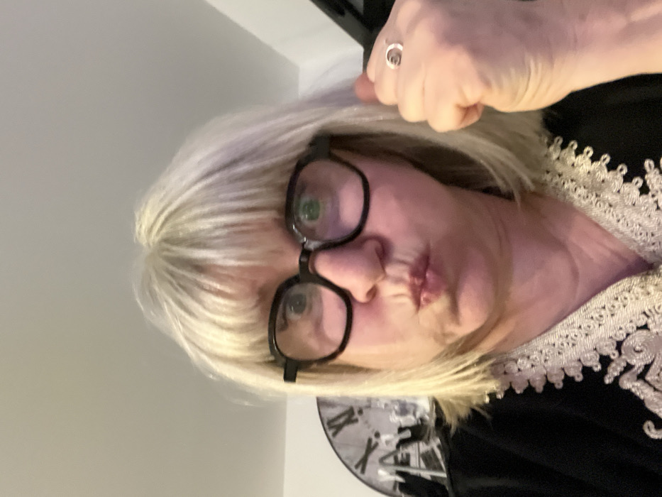 A selfie features a fair-skinned woman with short, blonde hair and black-framed glasses. Her lips are pursed in a slight pout. A ring adorns the index finger of her raised hand near her hair. She wears a black garment with intricate white lace trim along the neckline. In the background, a glimpse of a clock and other objects can be seen. The overall lighting appears soft and diffused.