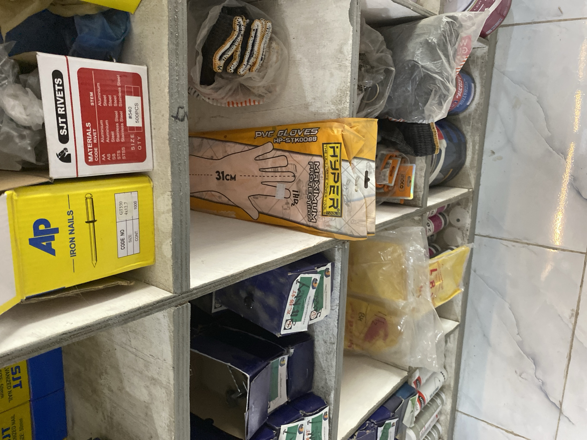The image shows a cluttered shelf in what appears to be a hardware store or workshop. The shelves are made of wood and contain various items including boxes of nails, rivets, gloves, and other hardware supplies. In the foreground, a yellow box with "Ap IRON NAILS" is visible. To the right of it, a box labeled "SJT RIVETS" is seen, with a table listing different materials and stem types. Below these, blue boxes with labels that are partially obscured by shadows are stacked. On the right side of the shelf, a package of "PVC GLOVES" is prominently displayed. Further to the right, a plastic bag contains several pairs of gloves. The overall environment appears to be dimly lit and disorganized, with items piled on top of each other. The flooring beneath the shelves consists of tiled surface.