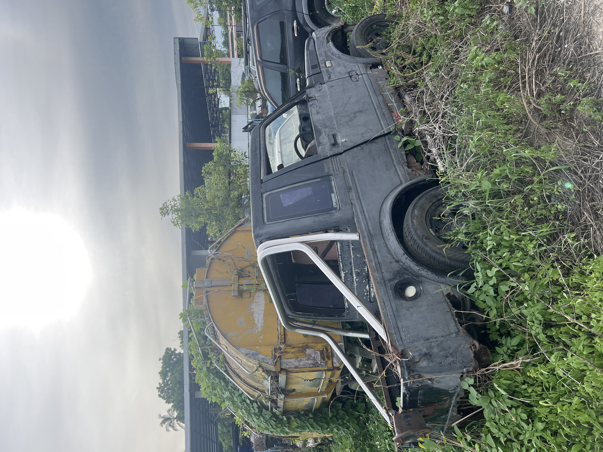 The image depicts a junkyard scene on Koh Phangan, Thailand. The primary subjects are several dilapidated vehicles, including a dark-colored SUV and a smaller, dark-colored utility vehicle, both overgrown with lush green vegetation. A large yellow cylindrical tank, also partially covered in vines, is visible behind the utility vehicle. The vehicles appear to be abandoned and in a state of disrepair, suggesting they are no longer in use.
The scene is set outdoors, with the overcast sky indicating a potentially cloudy or hazy day. The presence of dense foliage and vines strongly suggests a tropical or humid climate, consistent with the provided location context of Koh Phangan. The overall atmosphere is one of decay and neglect, with nature reclaiming the discarded objects.
Notable details include the rust and wear on the vehicles, the thick growth of plants, and the texture of the weathered yellow tank. There is no visible activity or event taking place; the image appears to be a static capture of the junkyard. The lighting is diffused due to the cloudy conditions, casting a soft, even light over the scene.
The text "GRIBBRI" is visible on the side of the smaller utility vehicle.