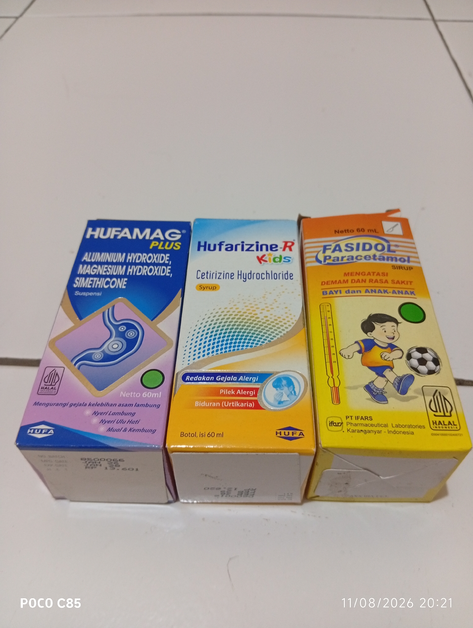 The image shows three cardboard boxes of medicine arranged in a line on a white surface. The box on the left is blue and purple and contains Hufamag Plus, an antacid suspension. The middle box is light blue and contains Hufarizine-R Kids syrup, an antihistamine for children. The box on the right is orange and yellow and contains Fasidol Paracetamol syrup for children. All three boxes are standing upright and facing the camera. The background is a plain white surface. The bottom of the image includes text identifying the device as "POCO C85" and a timestamp "11/08/2026 20:21." The items are likely on a table or floor. There are no people visible in the image. The lighting appears to be from overhead.
