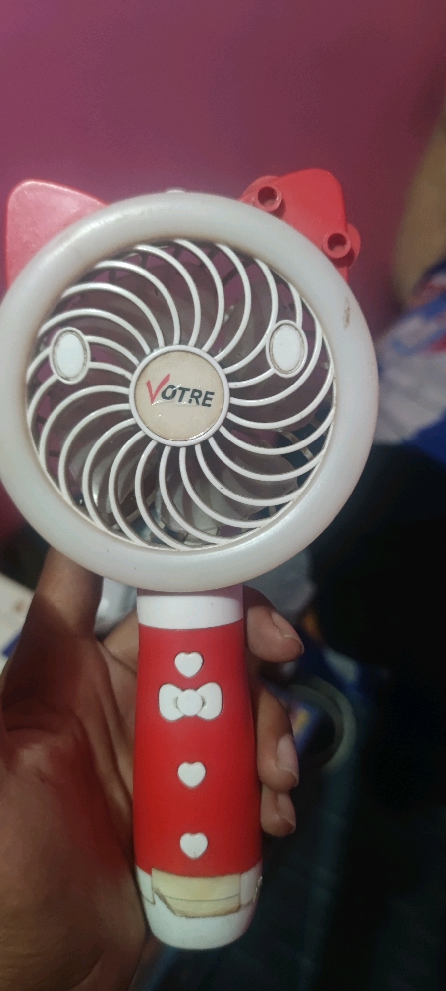 A handheld fan designed to resemble a Hello Kitty character is being held by a person's hand. The fan's head is white with red cat ears on top and features a circular fan grille with a "VOTRE" logo in the center. The handle is red with white heart and bow-shaped buttons. The background is out of focus and appears to be an indoor setting with a pink wall. The image is a close-up shot, showing the fan and the hand holding it. There is no information available about the specific location in Gambarsari, Indonesia.