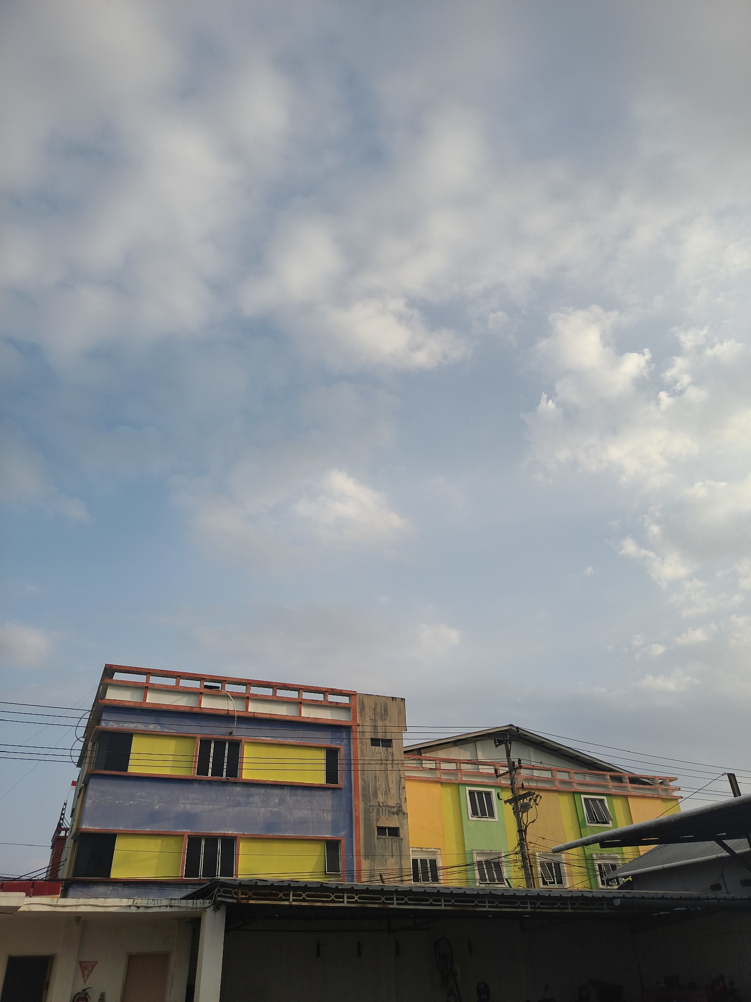 The image captures a view of a building in the city of Pangkalpinang, Indonesia. The building is multi-story and painted in vibrant colors, including blue, yellow, green, and orange. It has a modern architectural style with visible windows and balconies. Power lines are strung across the foreground, indicating an urban setting. The sky above is a clear blue with scattered white clouds. The image appears to be taken from a low angle, looking up at the building. There are no visible people in the frame.