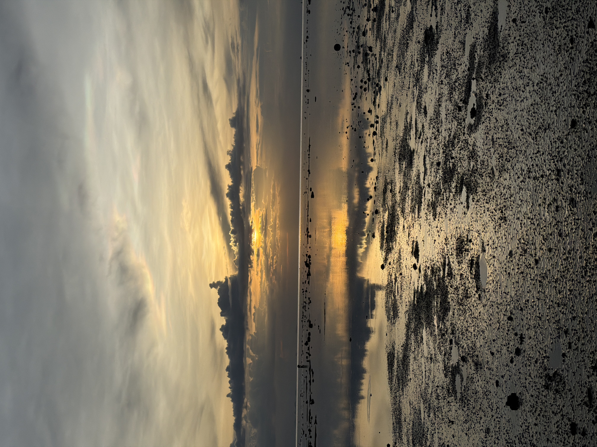 The image depicts a reflective, low-tide scene at sunset in Baan Plai Laem, Thailand. The sky is a gradient of grey and orange, with distinct cloud formations. The setting sun is partially obscured by clouds, casting a golden glow across the horizon and its reflection on the wet sand and shallow water. The foreground is characterized by a textured, uneven surface of dark sand interspersed with small rocks and puddles of water that mirror the sky. In the mid-ground, a solitary figure is visible standing in the shallow water, appearing to be engaged in observation or activity. The overall atmosphere is serene and natural, capturing a moment of transition between day and night on a coastal landscape.