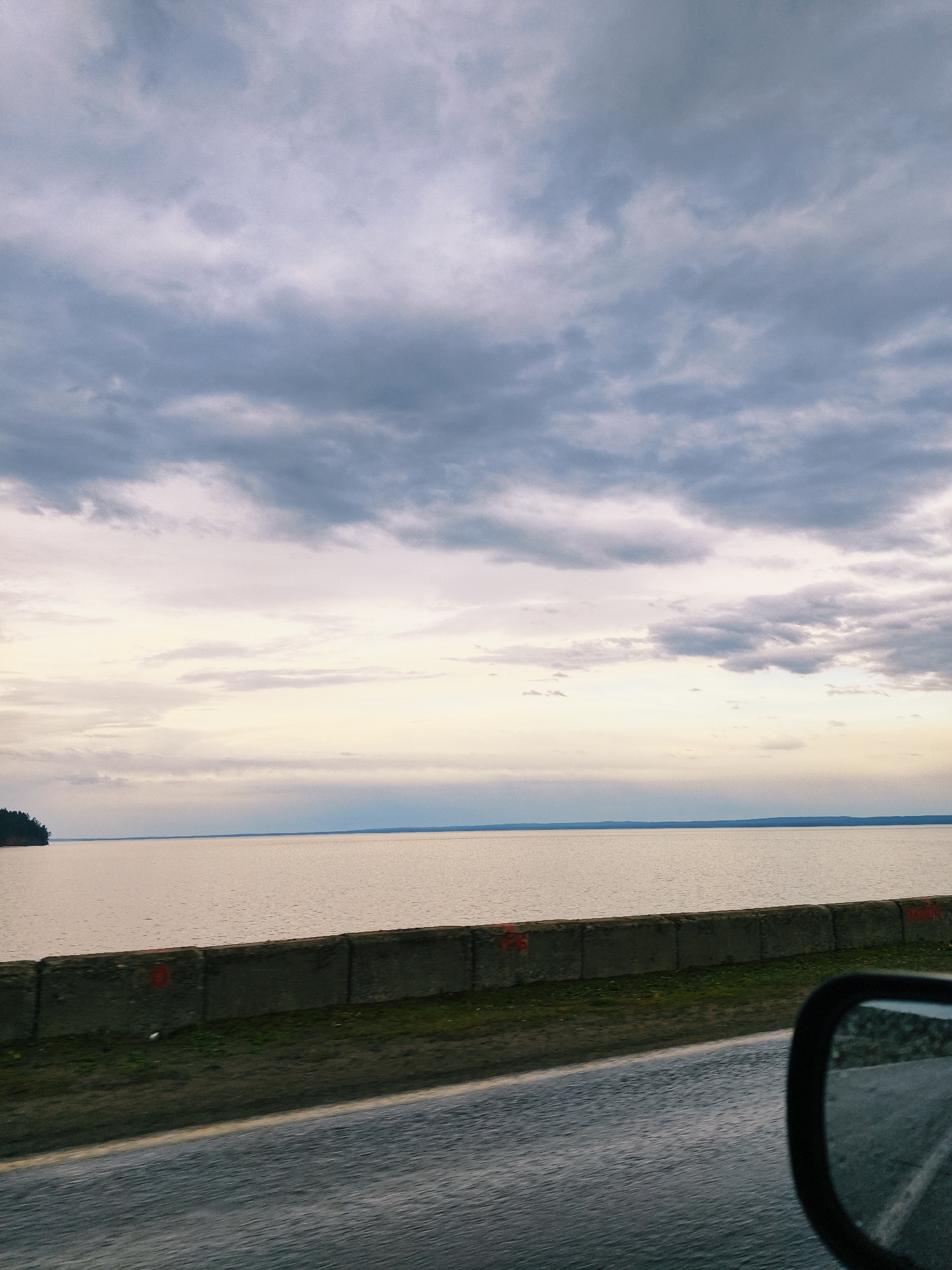 The image shows a view from a moving vehicle in the city of Chaikovsky, Russia. The camera is positioned at the road level, where the asphalt road fills the bottom part of the image. Next to the road is a concrete barrier with some minor vegetation growing along its base. Beyond the barrier is a large expanse of water, likely part of the Kama Reservoir, a defining feature of Chaikovsky.
Across the water, a distant shoreline with a line of trees can be seen. The sky is overcast with heavy, gray clouds covering most of the upper half of the image. The light is diffused, suggesting a cloudy day. The vehicle's side mirror is visible in the lower right corner of the image, partially reflecting the interior of the car. There are no people visible in the frame. The overall atmosphere is somber due to the overcast sky and the muted colors of the landscape.