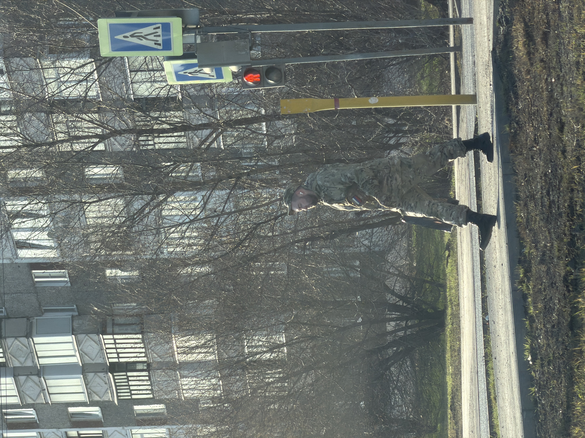 A person wearing camouflage clothing, a hat, and black boots is on the sidewalk next to a street. The person is near a pedestrian crossing sign and a traffic light showing a red light. Behind the person and the street infrastructure, there are bare trees and a multistory building with many windows. The photo was taken in the city of Izhevsk, Russia.