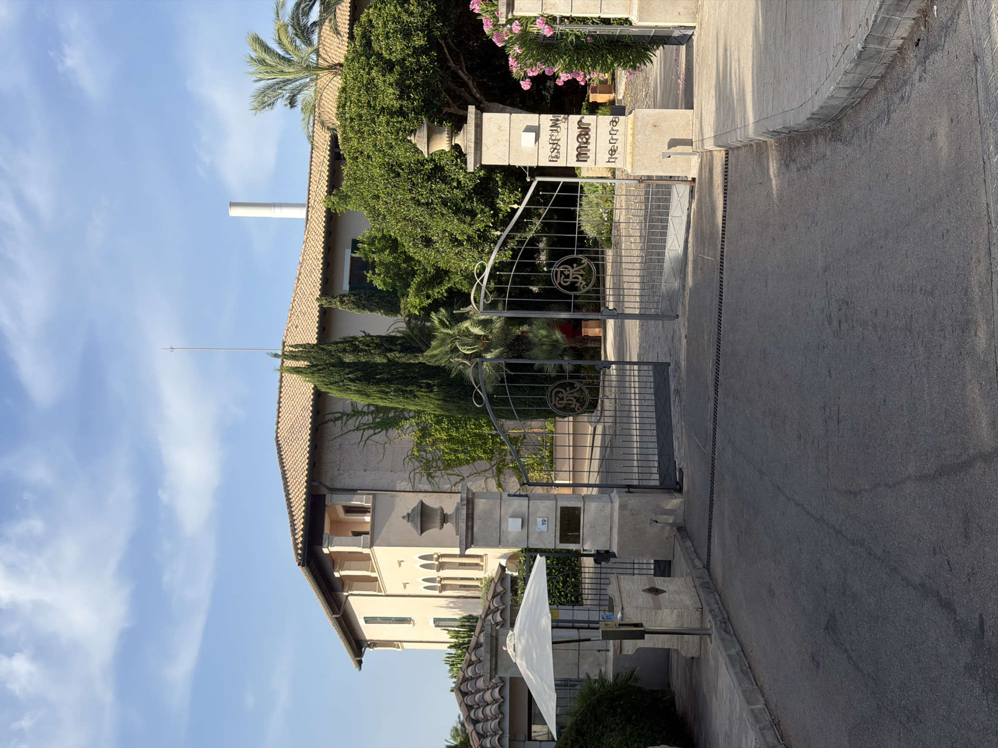 The image shows the entrance to a property with a large, ornate gate. The gate is open, revealing a paved driveway leading to a building. The building appears to be a large house or villa with a tiled roof and several windows. The property is surrounded by trees and vegetation. There is a sign on the gate that reads "ESTUDIO MAR HERRA". The location is identified as Calvià, Spain. The scene is set in daylight with a clear blue sky.