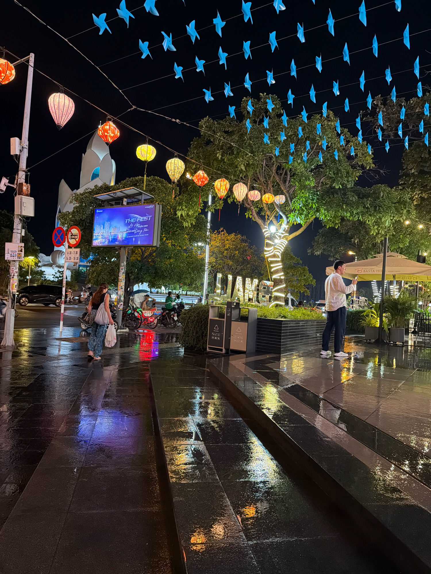 The image captures a nighttime scene in Nha Trang, Vietnam, likely a public square or pedestrian area, given the presence of decorative lights, a large screen, and street furniture. The ground is wet and reflective, indicating recent rain or cleaning, with colorful light reflections visible.
In the foreground, a person is walking away from the camera, carrying two white plastic bags. They are wearing a dark top and patterned pants. To the right, another person stands, facing away from the camera, looking at their phone and holding it up as if taking a photo or video. They are wearing a light-colored shirt and dark pants.
Overhead, strings of blue origami-like birds and traditional-style lanterns are hung, illuminated by the ambient light. A large tree in the middle ground is wrapped with strings of white lights, creating a festive atmosphere. A large digital screen displays "THE FEST" along with images of a city skyline. In the background, a modern white building with curved architectural features is visible, along with trees and outdoor seating areas of what appears to be a restaurant or cafe. Various signs and traffic signals are also present, suggesting a public thoroughfare. Motorcycles are parked on the side, a common sight in Vietnamese cities. The overall impression is one of a vibrant urban environment at night, decorated for a special event.