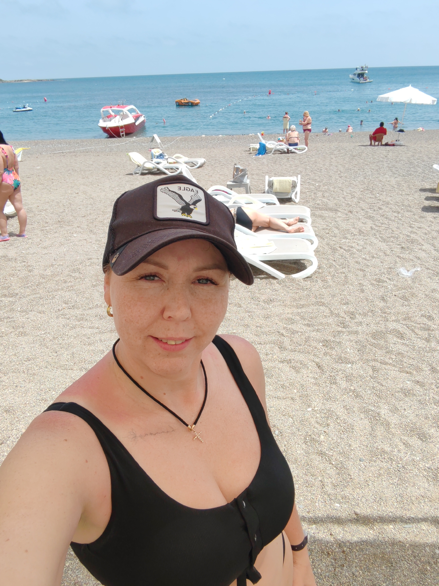 The image shows a woman in a black bathing suit and a brown baseball cap on a pebble beach. The cap has a patch with an eagle on it. She is wearing a gold earring and a black necklace with a gold cross. The background shows the Mediterranean Sea with several boats, including a red and white boat and another boat further out. Other people are visible on the beach, some lounging on chairs under a white umbrella, and others in the water. The sky is blue with some clouds. The location appears to be a beach in Alanya, Turkey.