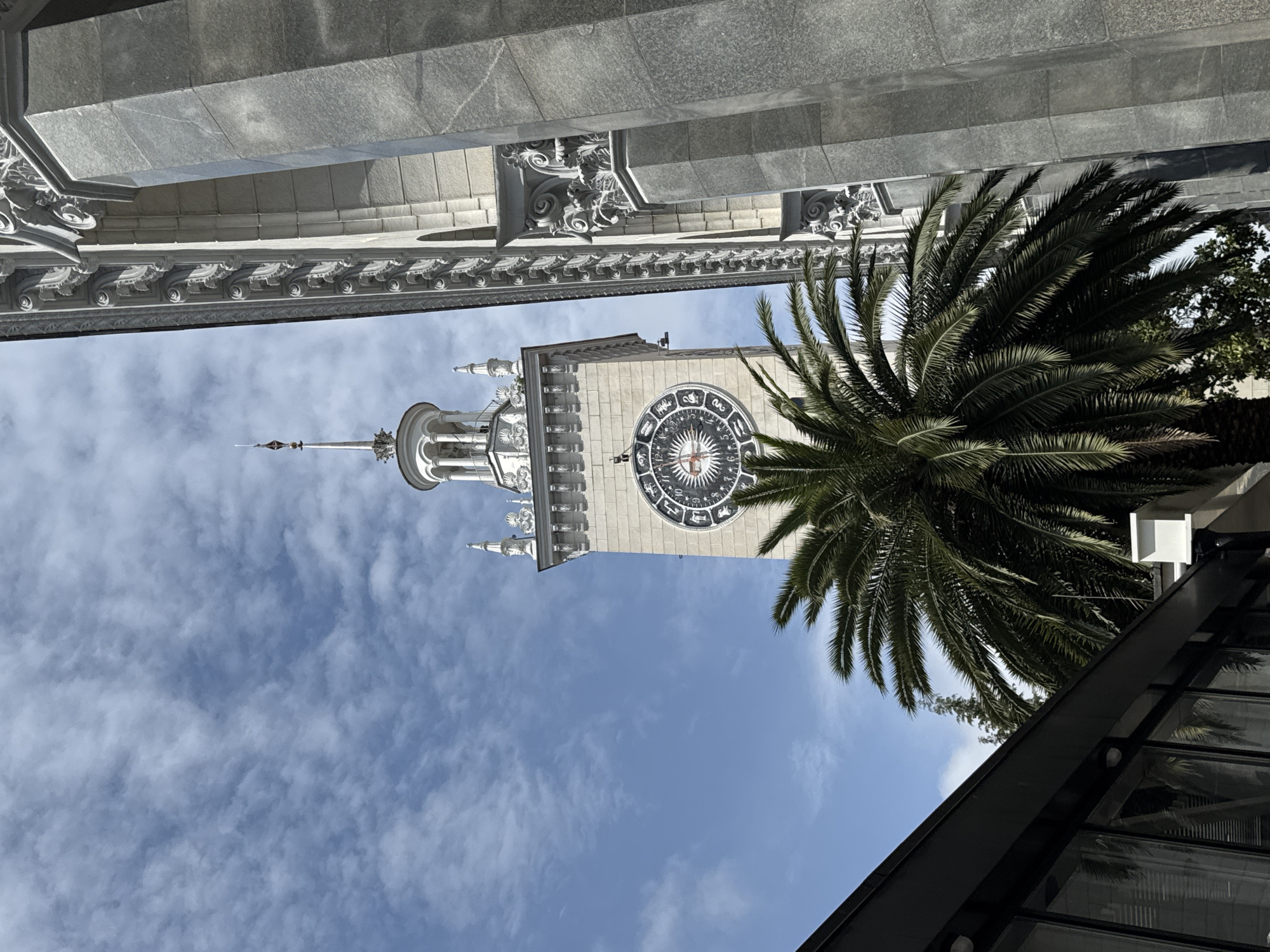 The image captures a view looking upwards towards the Sochi Marine Terminal clock tower. The clock tower is a prominent feature of the Sochi skyline. The clock face displays zodiac signs. A palm tree is visible to the right of the tower, partially obscuring the view. Architectural details of nearby buildings frame the shot at the top and bottom, suggesting the viewer is positioned close to these structures. The sky is blue with some clouds. The photograph suggests an outdoor setting in a sunny location.
