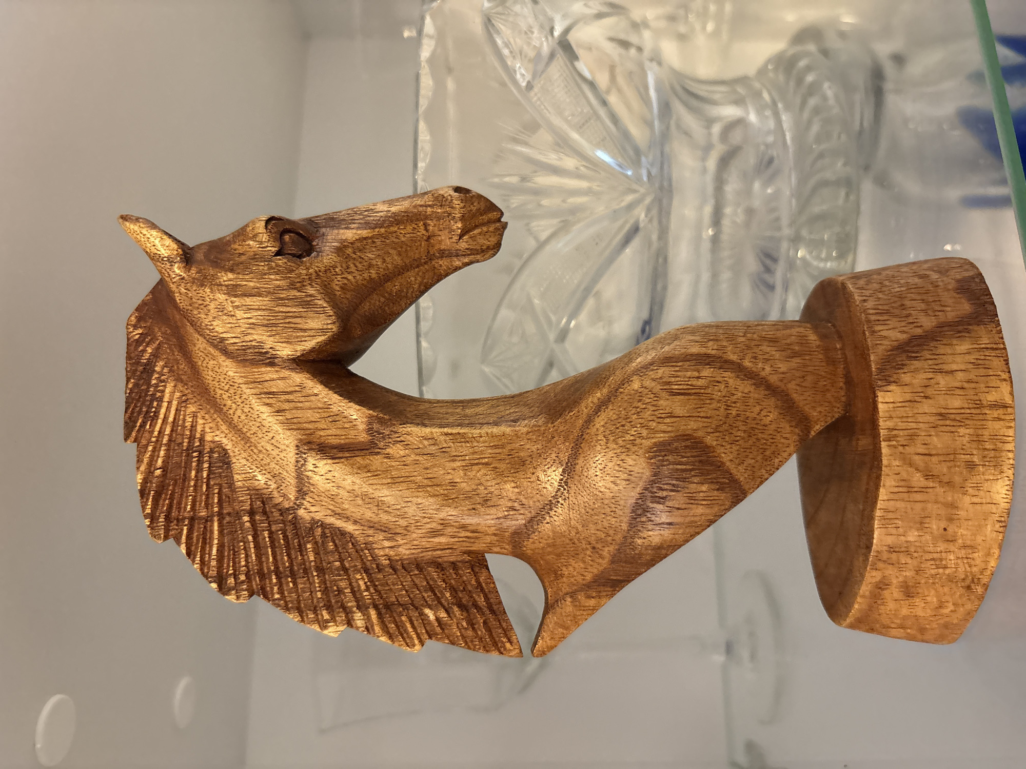 A close-up, eye-level shot captures a carved wooden horse head, the subject positioned centrally and slightly to the left. The horse head is crafted from a light brown wood, featuring textured details on the mane and a carved eye. It rests on a round wooden base. In the background, a clear crystal bowl with intricate patterns is visible, along with shelves and a white ceiling with two circular lights. This image was taken in Plovdiv, Bulgaria, specifically inside a home on the second floor, in the living room.