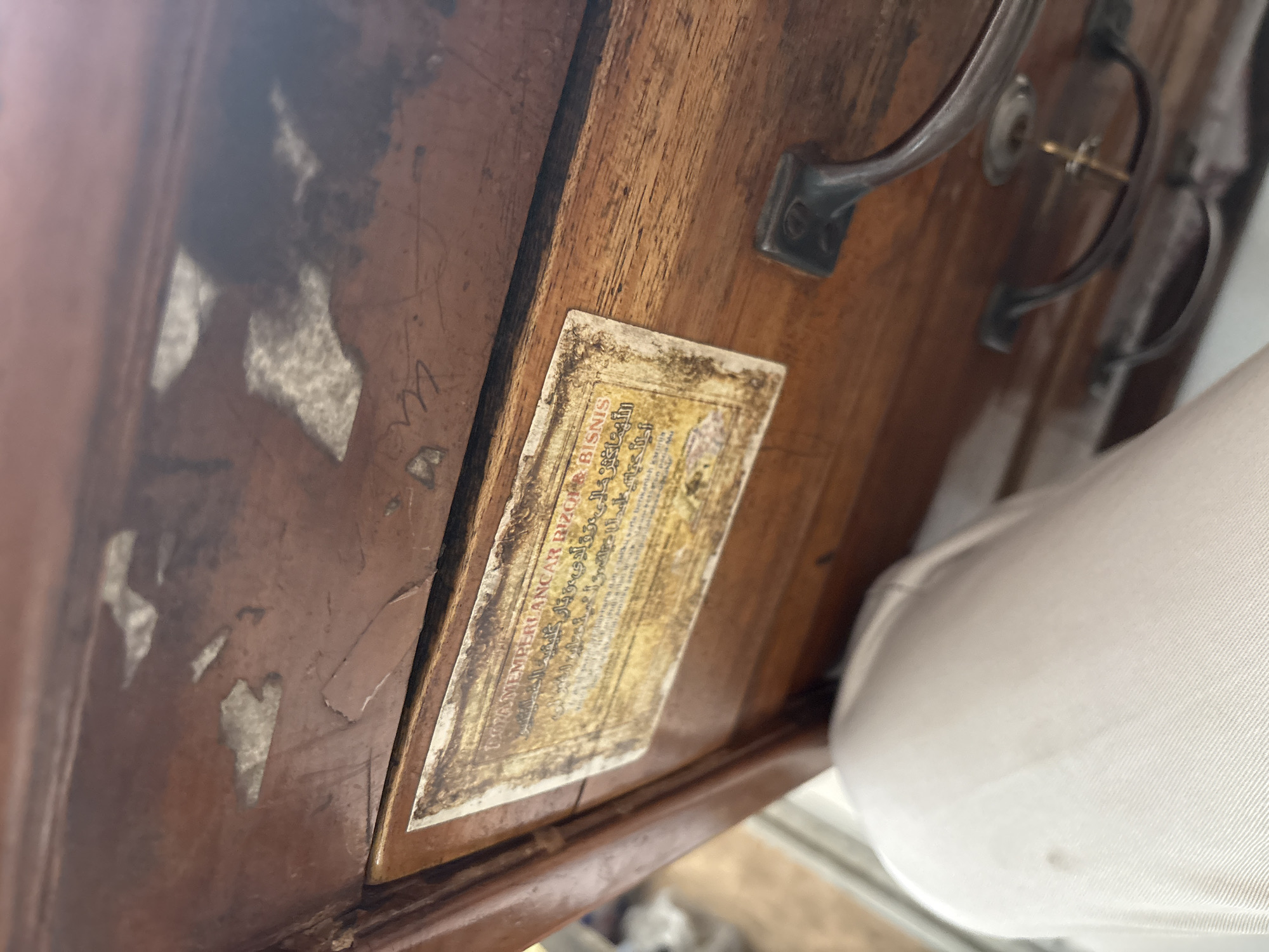 The image shows a close-up of an antique wooden drawer, possibly part of a desk or cabinet. The drawer has a distressed, worn finish with peeling varnish. A label with text, likely in Arabic script and possibly Malay or Indonesian, is affixed to the front of the drawer. The label is yellowed and aged. Two metal drawer pulls are visible, one on the upper portion of the drawer front, and another partially obscured by a white fabric item in the foreground. The lower pull has a small key inserted into its lock. The background is out of focus, but appears to be an indoor setting. The image is taken from a slightly elevated angle, looking down into the open drawer. The white fabric in the foreground suggests a person is seated or standing near the object, partially obscuring the lower part of the drawer. The object is likely located within a residential or commercial setting in Palimanan, Indonesia, given the context provided.