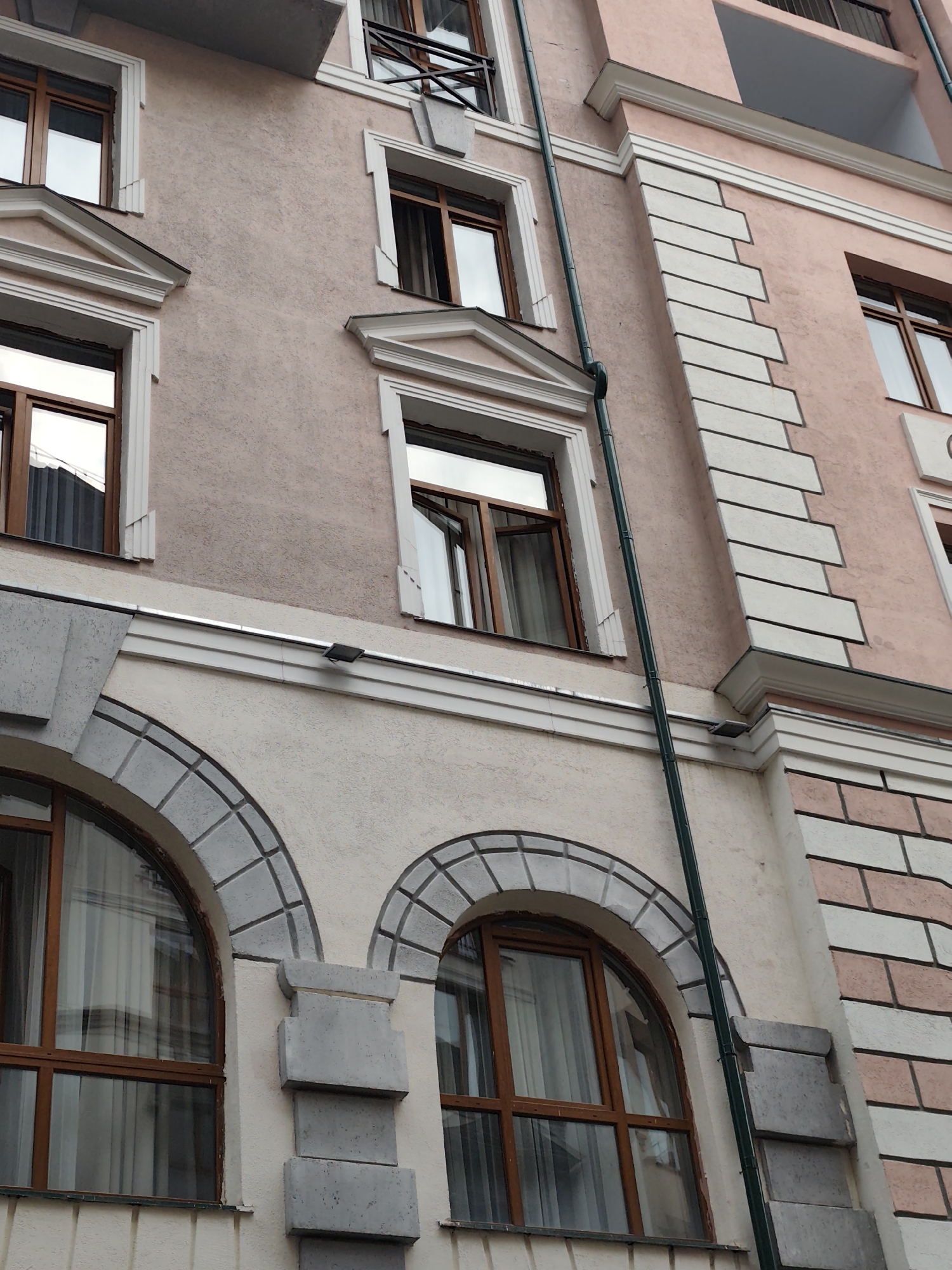 The image shows a low-angle view of a multi-story building with a pink facade in the city of Estosadok, Russia. The building features several windows with brown wooden frames. Some windows are rectangular, while others are arched. The facade is decorated with white architectural elements, including cornices and ornamental trim around the windows. A green drainpipe runs vertically along the building. The overall architectural style appears to be classical or neoclassical. The image is focused on the exterior of the building, with no people or specific activities visible.