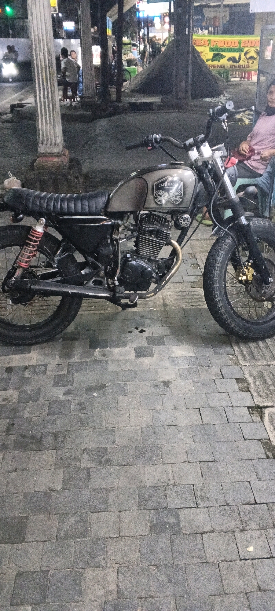 The image captures a street scene at night in the City of Medan, Indonesia, focusing on a customized cafe racer motorcycle parked on a stone-tiled pavement. The motorcycle is positioned centrally, viewed from the right side, revealing its metallic grey fuel tank with a "Cafe Racer" decal, black upholstered seat, exposed engine, and distinctive red rear shock absorbers. The front wheel, with its knobby tire and gold brake caliper, is visible on the right.
In the background, several elements suggest a commercial area. To the left, a white pillar with vertical grooves stands prominently. Further back, out of focus, are figures of people, some sitting and others standing, amidst what appears to be food stalls or shops illuminated by artificial lights. A yellow banner with text and images of seafood, including a crab and fish, is visible behind the motorcycle, indicating a restaurant or food vendor. The overall lighting is dim, characteristic of a nighttime urban environment, with the motorcycle and surrounding stalls illuminated by various light sources. The pavement itself shows signs of wear and some scattered dark spots, possibly oil stains.
