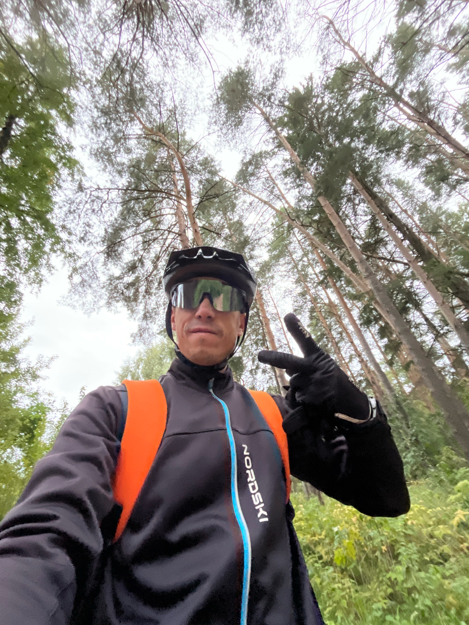 A low-angle, wide-shot selfie shows a person wearing a black cycling helmet, reflective sunglasses, a black jacket with an orange backpack, and black gloves. The person is outdoors, surrounded by tall trees with a cloudy sky overhead, suggesting a forest or park setting. The person is making a peace sign with their right hand. The jacket has the brand name "NordSki" in white letters with blue and white trim along the zipper. The background consists of a dense forest with thin, tall trees reaching towards the sky, characteristic of the coniferous forests found in the region of Chaikovsky, Russia, which is situated on the Kama River in the Perm Krai. The overall atmosphere is overcast and natural.