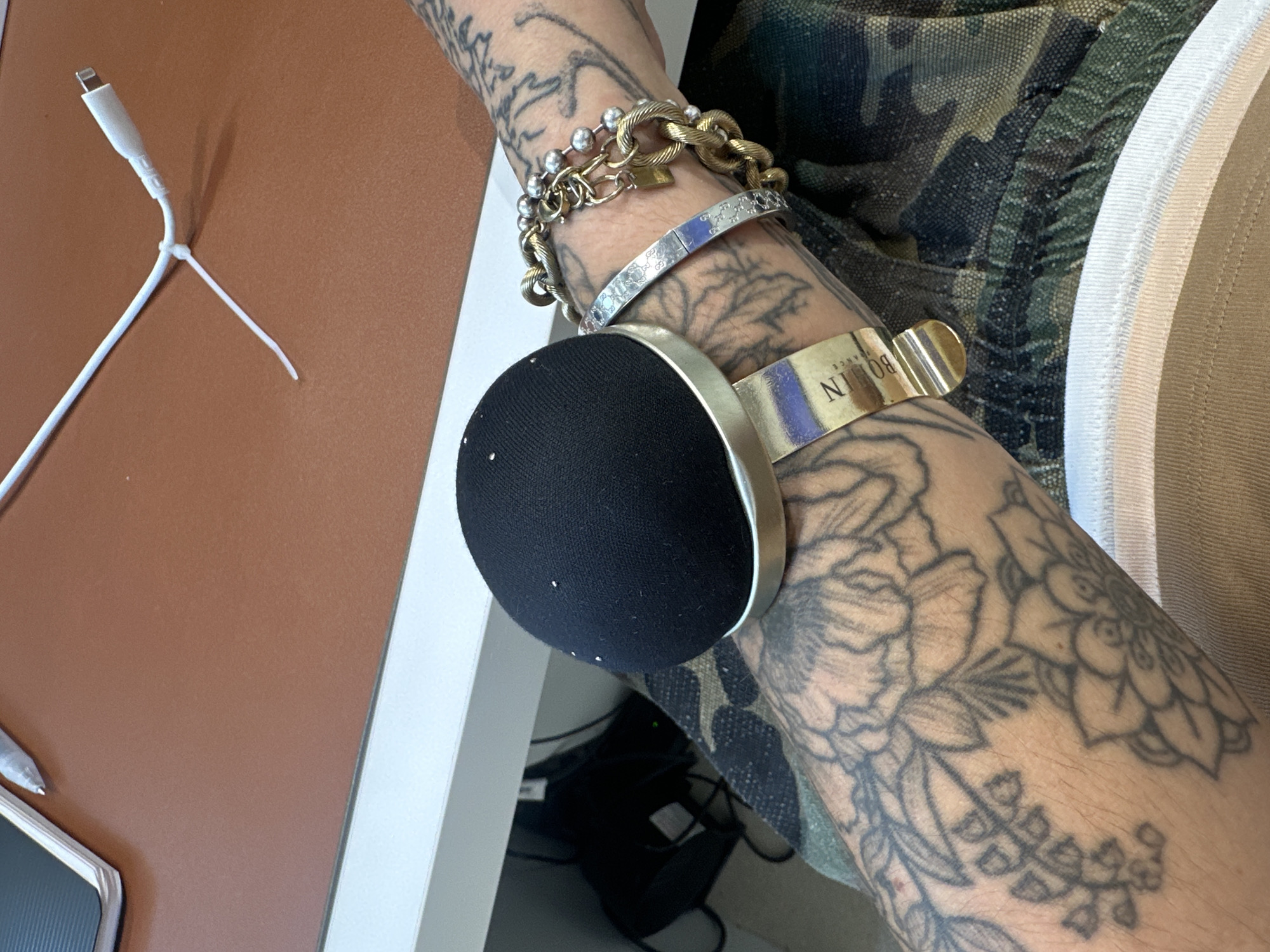 A close-up shot shows a tattooed arm resting on a desk or table. The arm has multiple bracelets. The uppermost is a gold and silver bracelet with interlocked chains. Below is a silver-colored bracelet featuring decorative etchings. A third bracelet is thick, gold in color, and inscribed with "BOIJN PARIS". An object that is round and black, possibly an electronic device, is partially visible near the wrist. The arm is tattooed extensively with intricate floral patterns. The person is wearing a camouflage garment with a white trim. The background includes a brown surface and a white cable. Beneath the table, dark objects and wires are seen.