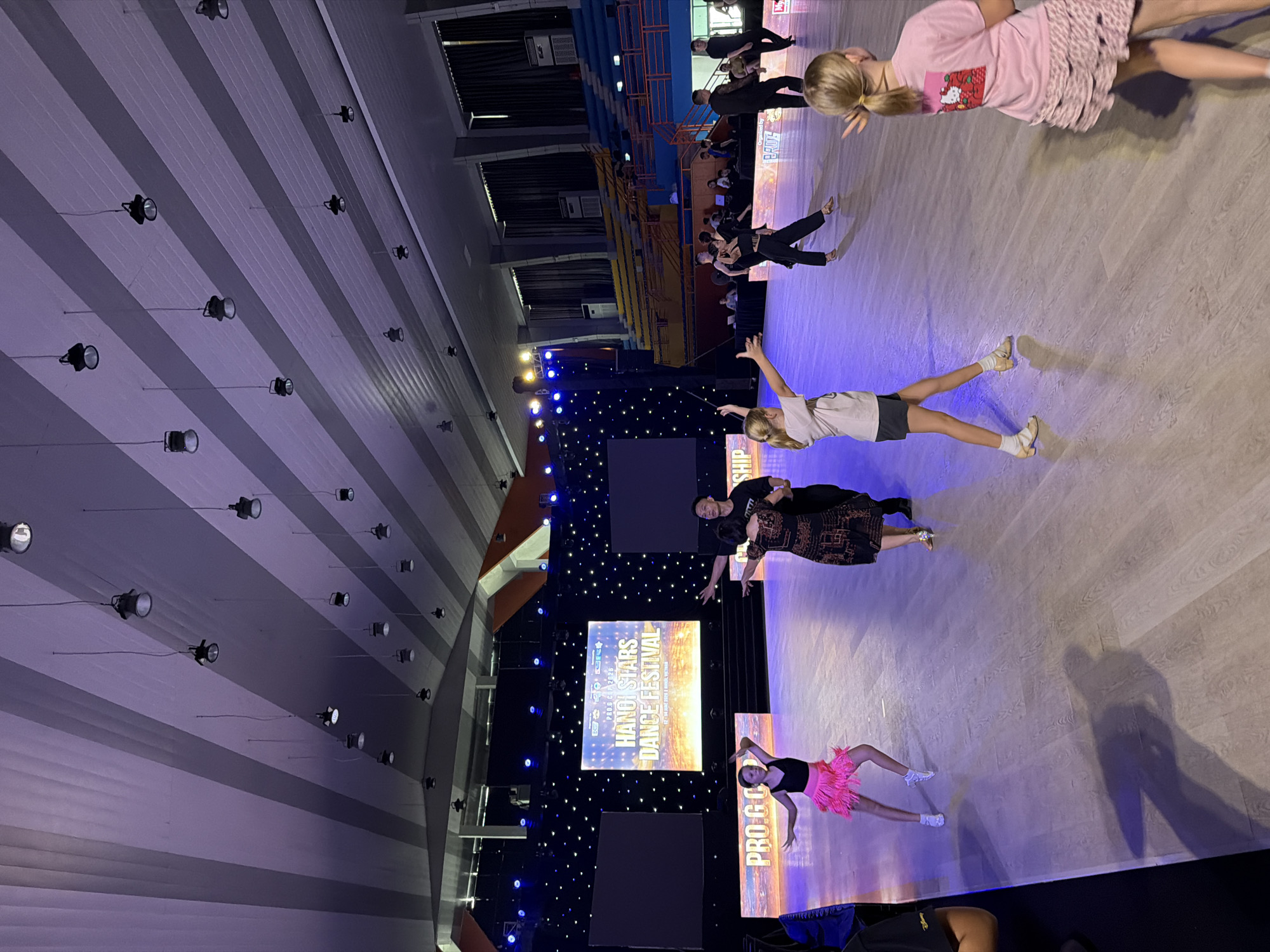 The image captures an indoor dance competition, likely the "Hanoi Stars Dance Festival" as indicated by the large screen display. The scene is set in a spacious hall with a polished wooden floor, illuminated by overhead spotlights and stage lighting that casts purple and blue hues. In the background, tiered seating areas are visible, some occupied by spectators. Several individuals, both children and adults, are engaged in dancing across the floor, dressed in performance attire. A group of dancers in black is positioned towards the right, while a young girl in a pink fringe skirt is in the foreground on the left. The overall atmosphere suggests a formal dance event.