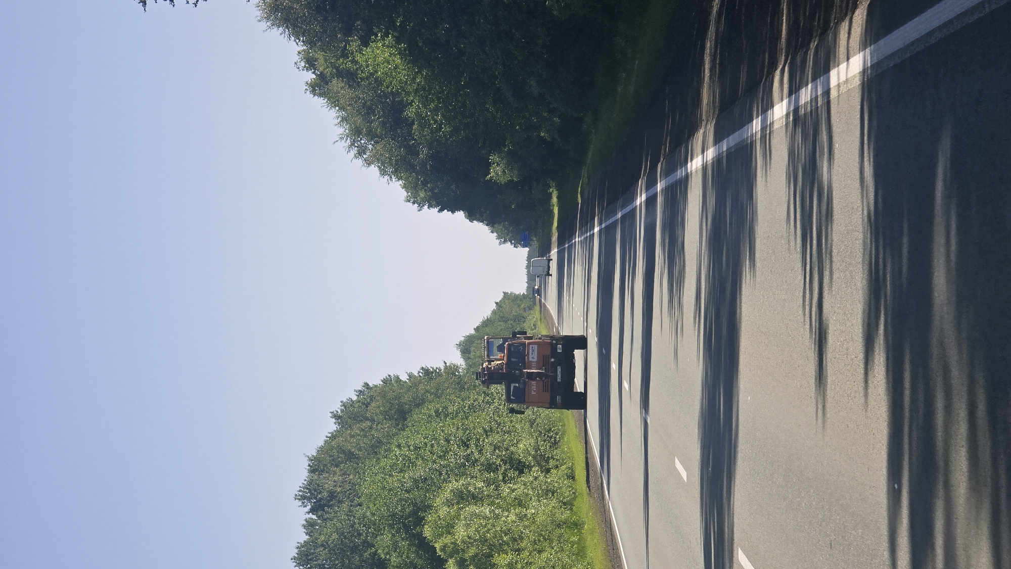 An orange truck with a crane attachment is driving down a two-lane road lined with dense green trees on both sides. The sky is clear and blue. There are other vehicles visible in the distance, including a white van. The road has white lane markings and the sun is casting shadows from the trees onto the asphalt. The scene appears to be on a sunny day in a rural or suburban area.