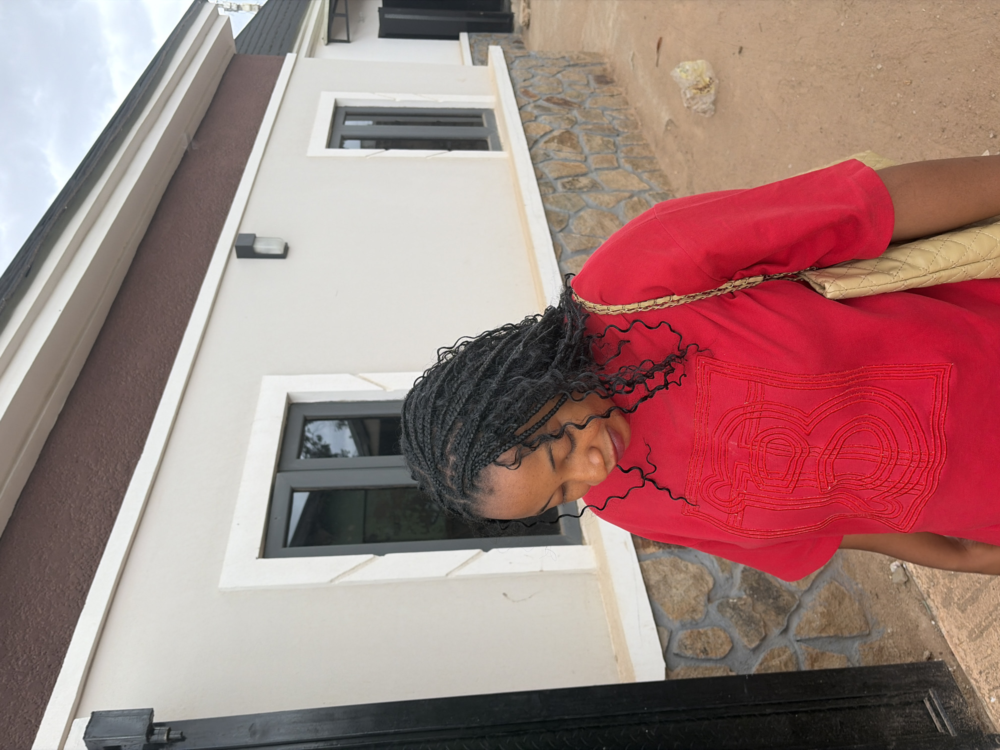 An outdoor, slightly rotated image captures a person standing next to a building in what appears to be a residential area in Jos, Nigeria. The main subject is a young woman, positioned on the right side of the frame, looking downwards with a calm or contemplative expression. She has dark, small box braids that frame her face and fall around her shoulders. She is wearing a bright red short-sleeved top, which features an elaborate, interwoven, possibly monogram-like design embroidered in red thread on the chest, resembling stylized "BB" or similar overlapping characters. A light beige or cream-colored quilted shoulder bag with a thin gold chain strap rests on her right shoulder.
The background consists of a modern-looking building with light off-white or beige walls. The lower section of the building's facade is clad with irregular natural stones in shades of grey and brown, adding a rustic texture. Dark gray-framed windows with visible glass reflect parts of the sky and surroundings. A dark gray outdoor light fixture is mounted on the wall. The ground is unpaved, consisting of light brown dirt or sand with scattered small stones. The sky in the upper left corner is overcast, indicating a cloudy daytime.