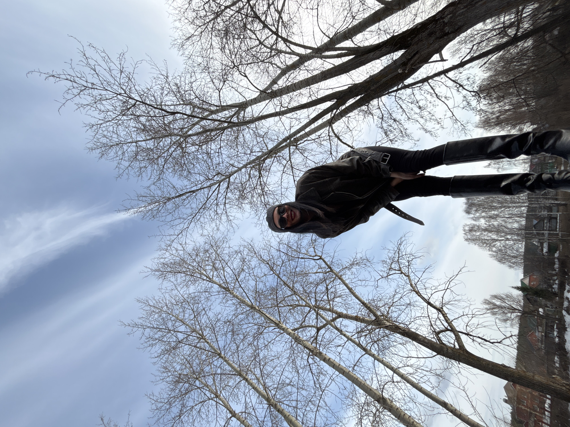 A full-length photograph depicts an individual standing outdoors in what appears to be a park-like setting. The individual is wearing a dark jacket, dark pants, and dark boots, possibly leather. They are also wearing sunglasses. The photograph is taken from a low angle, looking up towards the person and the sky.
Behind the individual, there are bare trees reaching up toward a sky with some light clouds. In the distance, across what might be a body of water, there are buildings, some of which appear to be residential. Based on the architectural style and the geographical reference, the location is Chaikovsky, Russia. The overall tone of the image is somewhat subdued, possibly taken on a cool or overcast day.