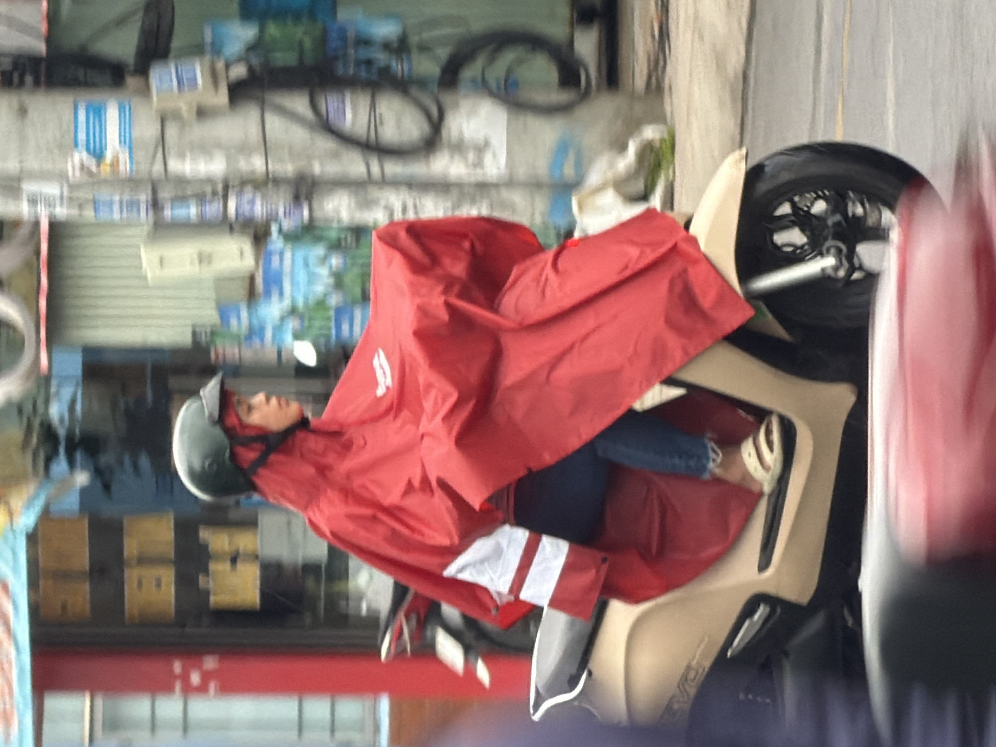 A person wearing a red poncho and a dark green helmet rides a tan scooter on a paved road. The person's face is partially obscured by the helmet and poncho, but they appear to be looking forward. The scooter has a black seat and the front wheel is visible. The background shows a building with yellow boxes on shelves and a utility pole with wires and signs. The scene is consistent with a street in Nha Trang, Vietnam.