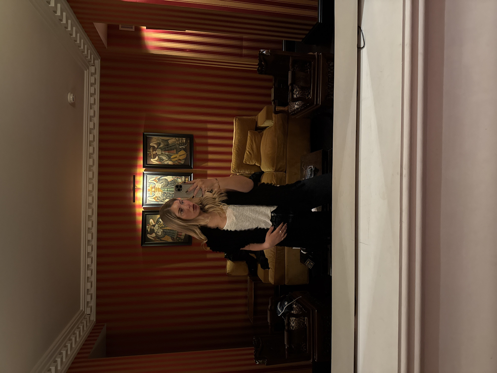 A fair-skinned, blonde-haired woman stands in front of a mirror in a room with reddish-brown and gold striped wallpaper. The room features ornate gold-framed artwork on the wall. A gold-colored couch and dark ornate wooden furniture are present. The location is possibly Paris, France, given the style of the architecture and interior design. The woman appears to be taking a photo of herself in the mirror with her phone. She is wearing a white top, a black jacket, and black pants. The mirror is framed by a white border.