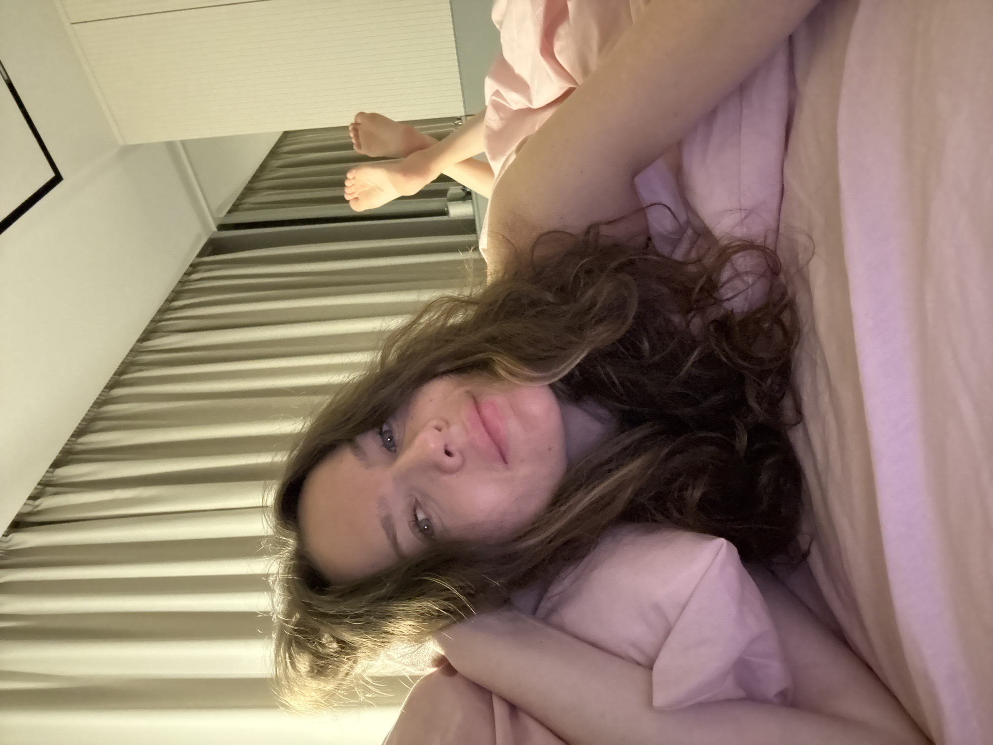 A woman lies on a bed, propped up on pillows. She is wearing a pink garment and has long, curly brown hair. Her eyes are open and she is looking at the camera. Her feet are visible crossed over each other in the background. The room appears to be a bedroom, with curtains and a closet visible. The lighting is dim.