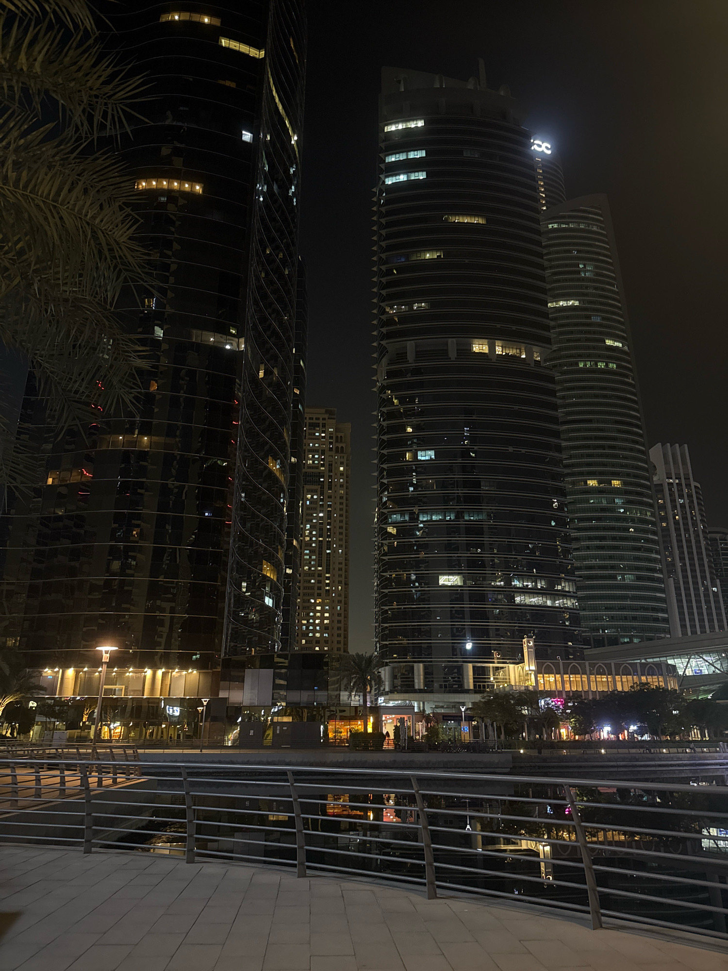 This image depicts a night-time scene of a waterfront promenade in Dubai, United Arab Emirates, likely in the Dubai Marina area. Dominating the background are several modern skyscrapers, characterized by their dark, reflective facades illuminated by interior lights. Two prominent, cylindrical towers with ribbed exteriors stand side-by-side, with one featuring a glowing white logo at its apex. To the left, another building with a dark, glossy exterior and spiraling architectural details is partially obscured by palm fronds. Further in the distance, a much taller, rectangular building displays a grid of numerous lit windows.
In the foreground, a paved walkway with a stone pattern leads towards a metal railing that separates it from a dark body of water. The water reflects the lights of the buildings and the promenade. Along the waterfront, trees and low-rise structures are visible, also illuminated by streetlights and interior lights, suggesting commercial or residential spaces. The overall atmosphere is one of urban sophistication and nighttime activity, with the city lights creating a dramatic contrast against the dark sky. There are no discernible people in the image.