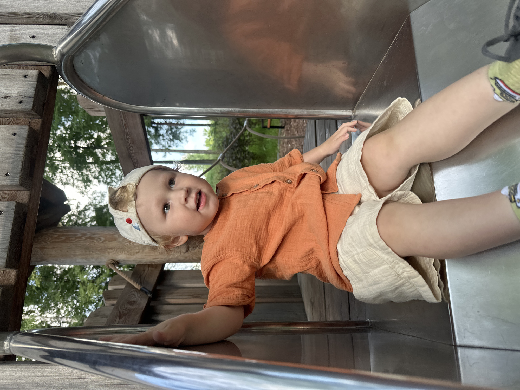 A young child wearing an orange shirt, cream-colored shorts, and a baseball cap is sitting on a metal slide at a playground. The playground appears to be made of wood and metal and is surrounded by trees. The child is holding onto the side of the slide and looking to the right. The image was taken in Votkinsk, Russia.