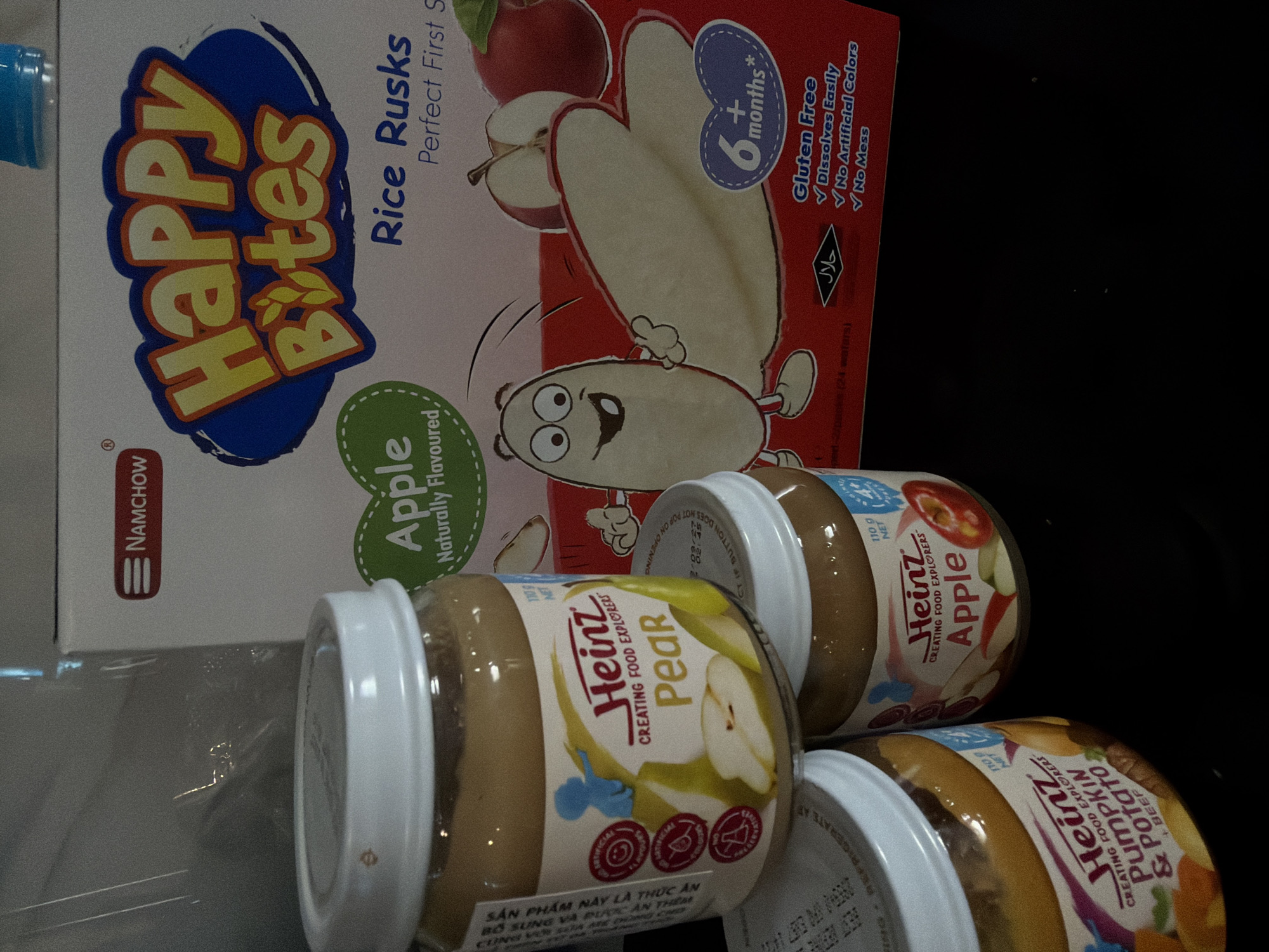 The image shows a box of "Happy Bites" rice rusks, labeled "Apple Naturally Flavoured," positioned behind three jars of Heinz baby food, namely Pear, Apple, and Pumpkin & Potato. The rusks are marked as suitable for babies 6+ months and are gluten-free. The Heinz jars are labeled "Creating Food Explorers." The scene appears to be in Nha Trang Ward, Vietnam, based on the Vietnamese text on the Heinz pear jar. The image appears to be taken indoors.