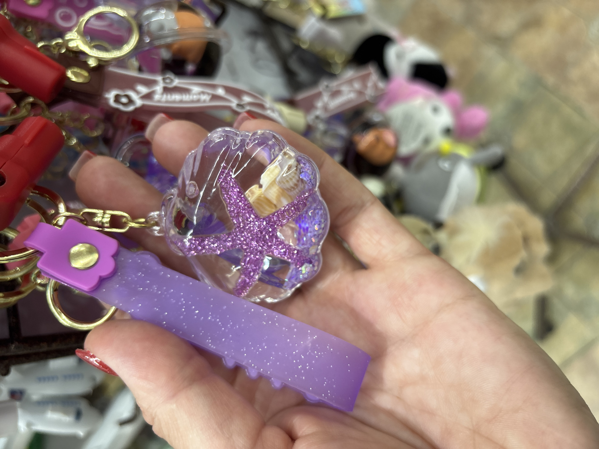 A close-up shot shows a person's hand holding several colorful keychain charms. The hand has red nail polish on one finger. The charms include a clear, shell-shaped trinket filled with tiny seashells and purple glitter, with a purple starfish inside. Another charm is a flat, rectangular piece of translucent purple plastic with glitter. A purple flower-shaped charm with a gold circle in the center is attached to a gold chain, along with a red object that appears to be a small plastic nozzle or spray bottle. The background is blurry but suggests a display of various trinkets and small toys, possibly in a store. Some of these background items include plush toys, one with pink ears and a grey body, and other small accessories. The lighting is bright, indicating an indoor setting.
