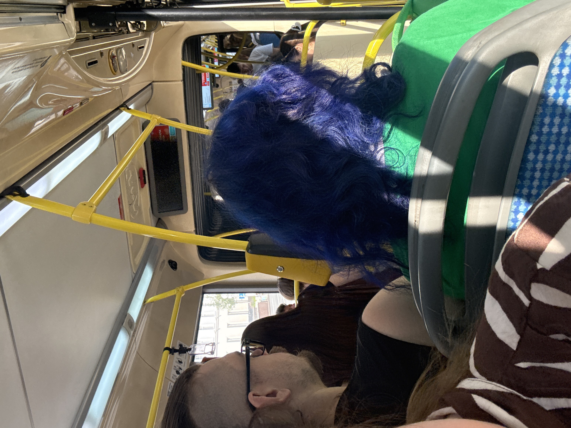 The image shows the interior of a public bus in Moscow, Russia, with passengers. In the foreground, a person with vibrant blue hair, wearing a green garment, is seated. To the left, another passenger with dark hair and glasses is visible. Yellow poles and handrails, characteristic of bus interiors, are prominent throughout the scene. The windows offer a glimpse of a street outside, with buildings and trees suggesting an urban environment. The lighting indicates it is daytime.