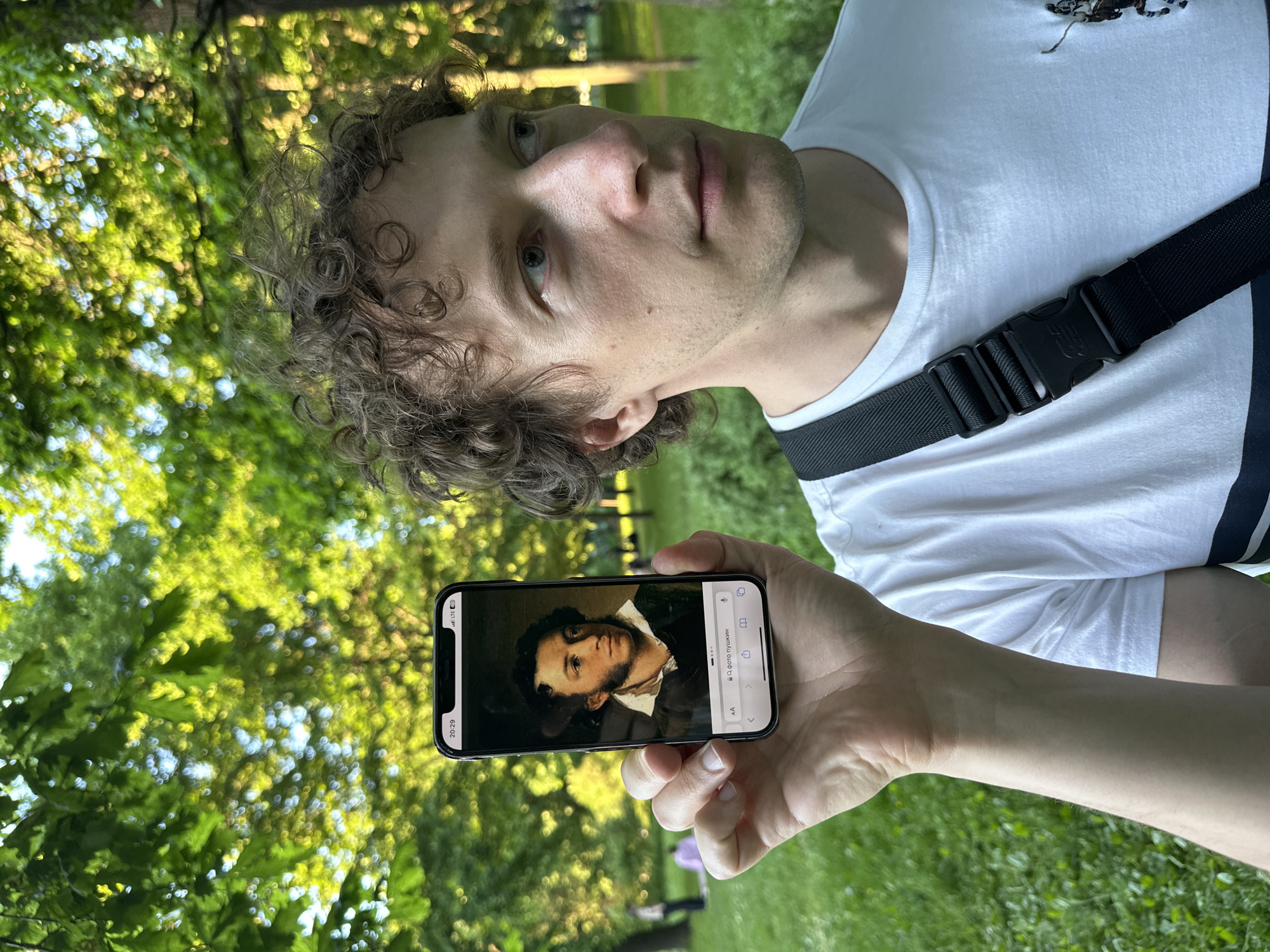 A male individual with fair skin, curly brown hair, and blue eyes, appearing to be in his late twenties to early thirties, is depicted in an outdoor setting. He is wearing a white t-shirt with a horizontal navy stripe across the lower torso and a black cross-body bag with a plastic buckle. He holds a black iPhone vertically in his left hand, presenting its screen towards the viewer. The phone's screen displays a portrait painting of Alexander Pushkin. The top left of the screen shows "20:29" and the top right indicates "LTE" signal and battery status. A search bar at the bottom of the phone screen contains the Cyrillic text "Q фото пушкин" (Q photo pushkin). The man is looking towards the right side of the frame, past the camera. The background consists of a public park in the city of Saint Petersburg, Russia, characterized by lush green trees with dappled sunlight creating bokeh effects, and green grass. Several blurred figures are visible in the distant background, suggesting other park visitors.